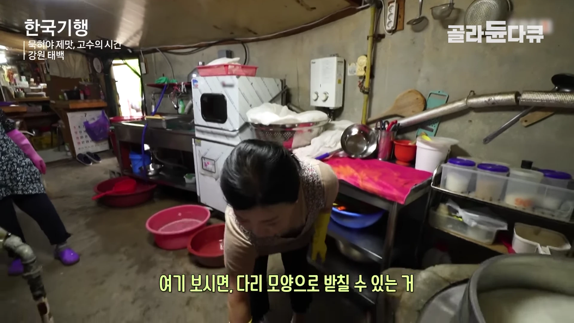주방에서 앞치마와 고무장갑을 착용한 여성이 허리를 숙여 무언가를 보여주고 있으며, 주변에는 낡은 주방 도구들과 조리대, 다양한 크기의 대야와 용기들이 놓여 있습니다.