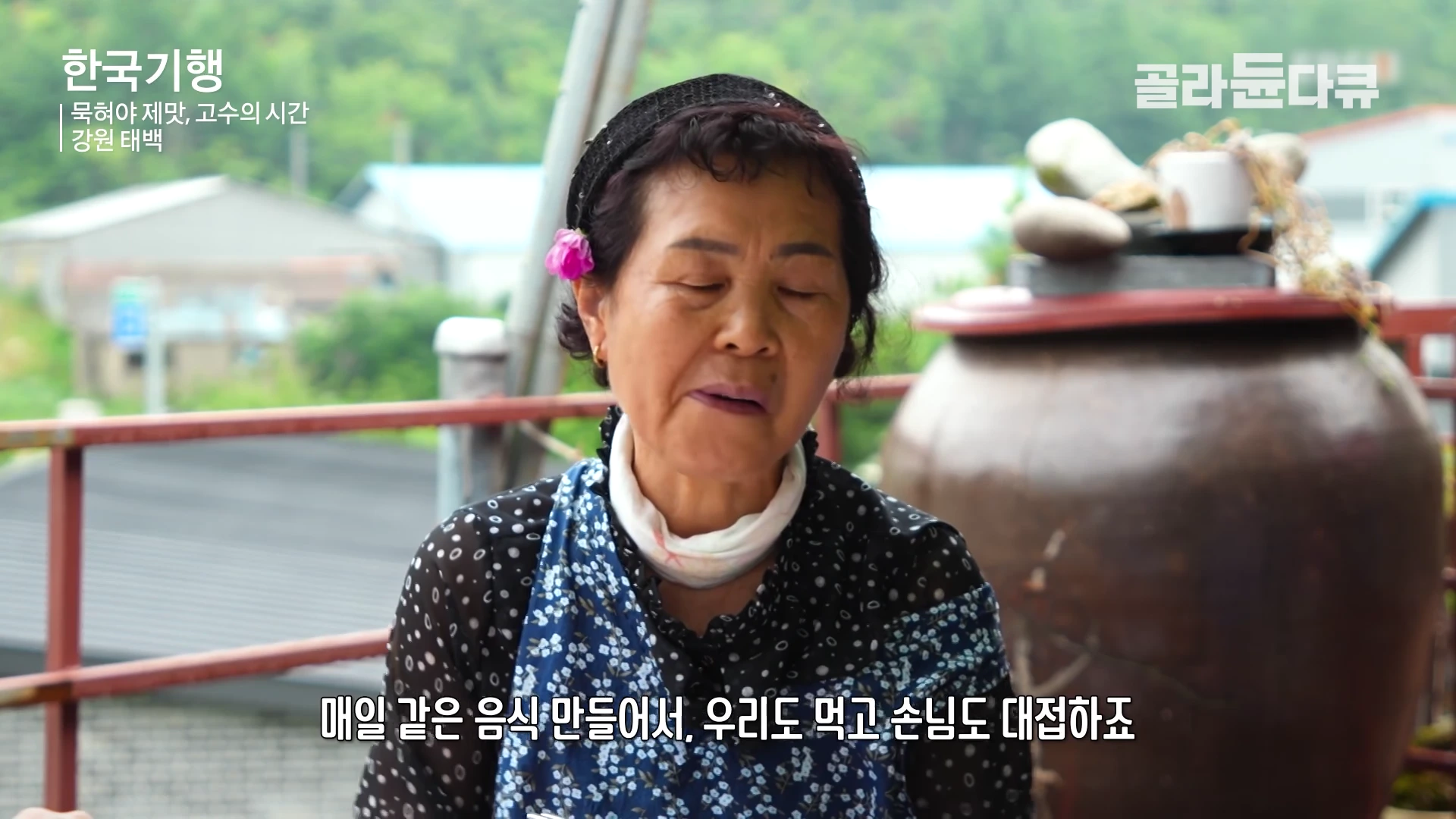 검은 머리띠를 하고 꽃 장식을 단 중년 여성이 야외에서 인터뷰하고 있는 모습