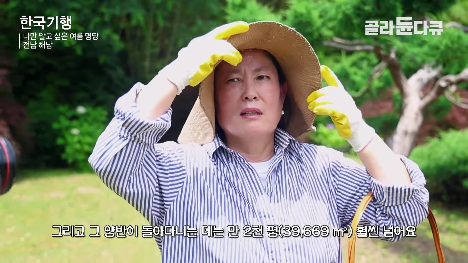 밀짚모자를 쓰고 줄무늬 셔츠를 입은 중년 여성이 야외 정원에서 노란 작업용 장갑을 낀 채 손으로 모자 챙을 잡고 어딘가를 바라보고 있는 모습.