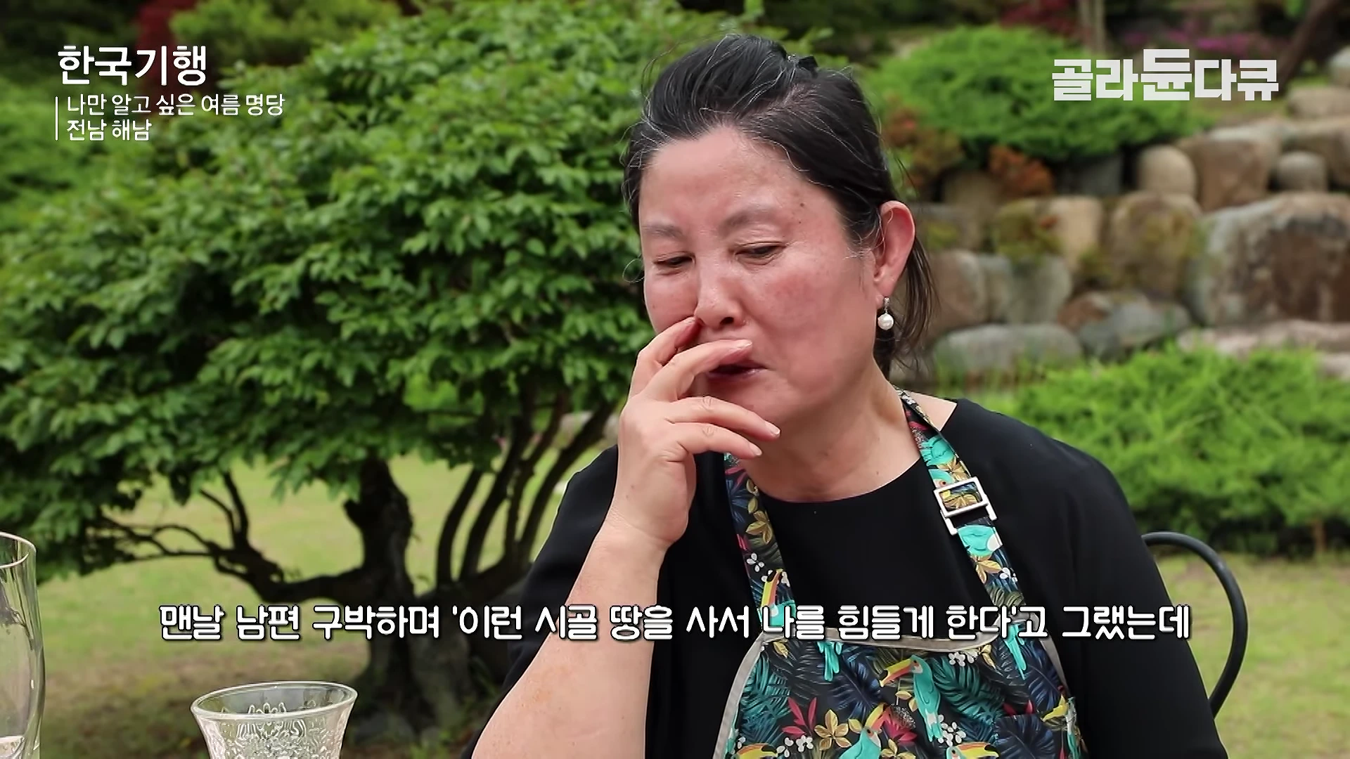 푸른 정원을 배경으로 검은색 상의를 입은 중년 여성이 고민하는 듯한 표정으로 앉아 있는 모습.