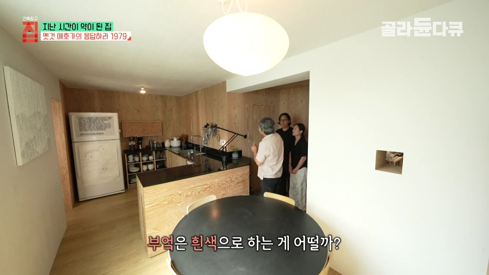 합판 벽과 흰색 천장이 조화를 이루는 주방 공간에서 세 사람이 대화를 나누고 있는 모습