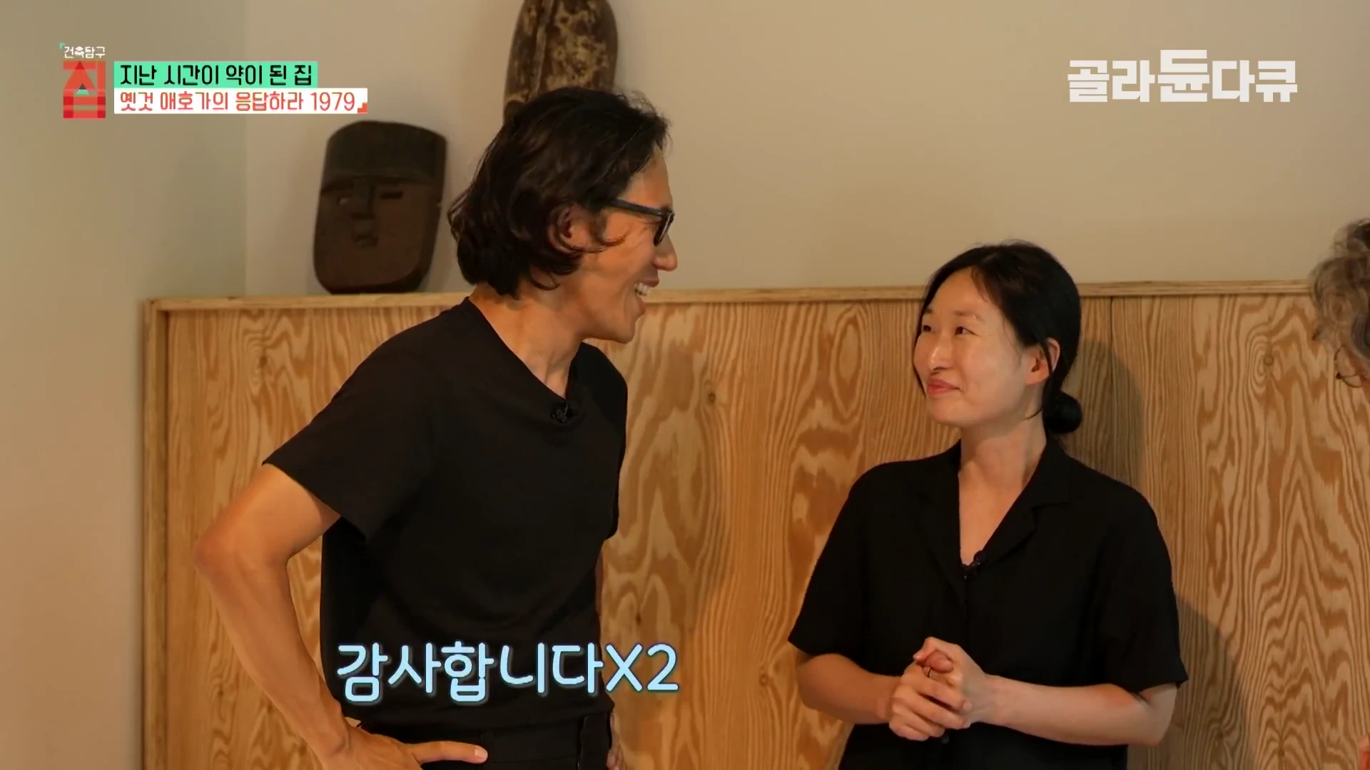 실내에서 검은색 상의를 입은 남녀가 서로를 바라보며 이야기를 나누고 있다. 배경에는 나뭇결이 드러나는 목재 벽면과 민속 가면 장식이 있다.