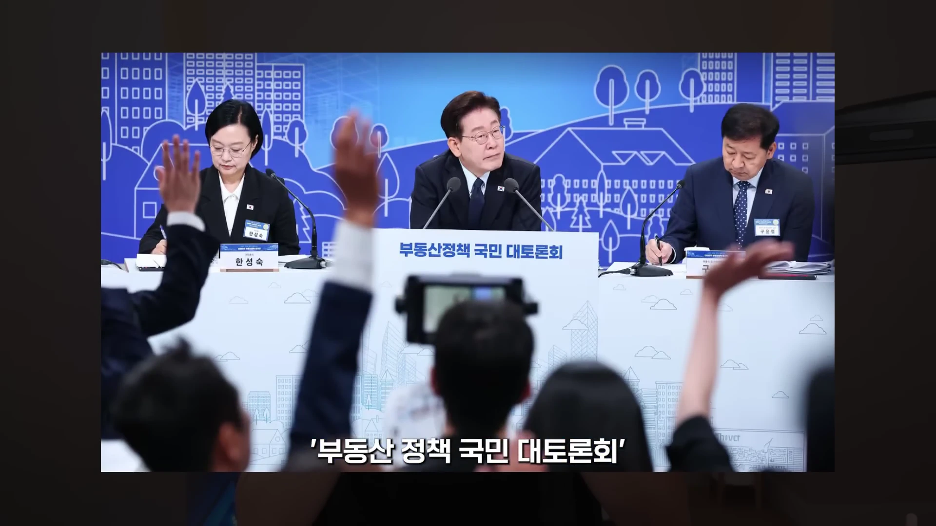 푸른색 배경의 무대에서 세 명의 인물이 앉아 토론을 진행하고 있으며, 앞쪽에는 손을 든 청중들의 모습이 보이는 영상 장면입니다.