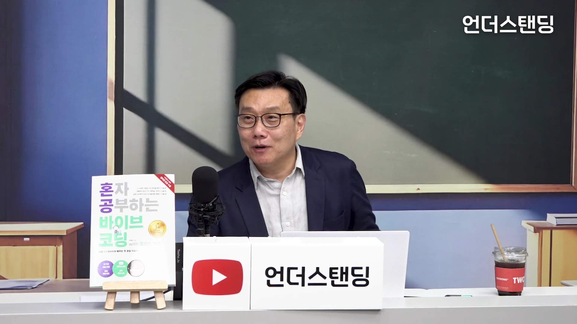 안경을 쓴 남성 출연자가 책상 앞에 앉아 인터뷰를 진행하고 있으며 책상 위에는 '혼자 공부하는 바이브 코팅'이라는 제목의 책과 마이크, 커피잔이 놓여 있습니다.