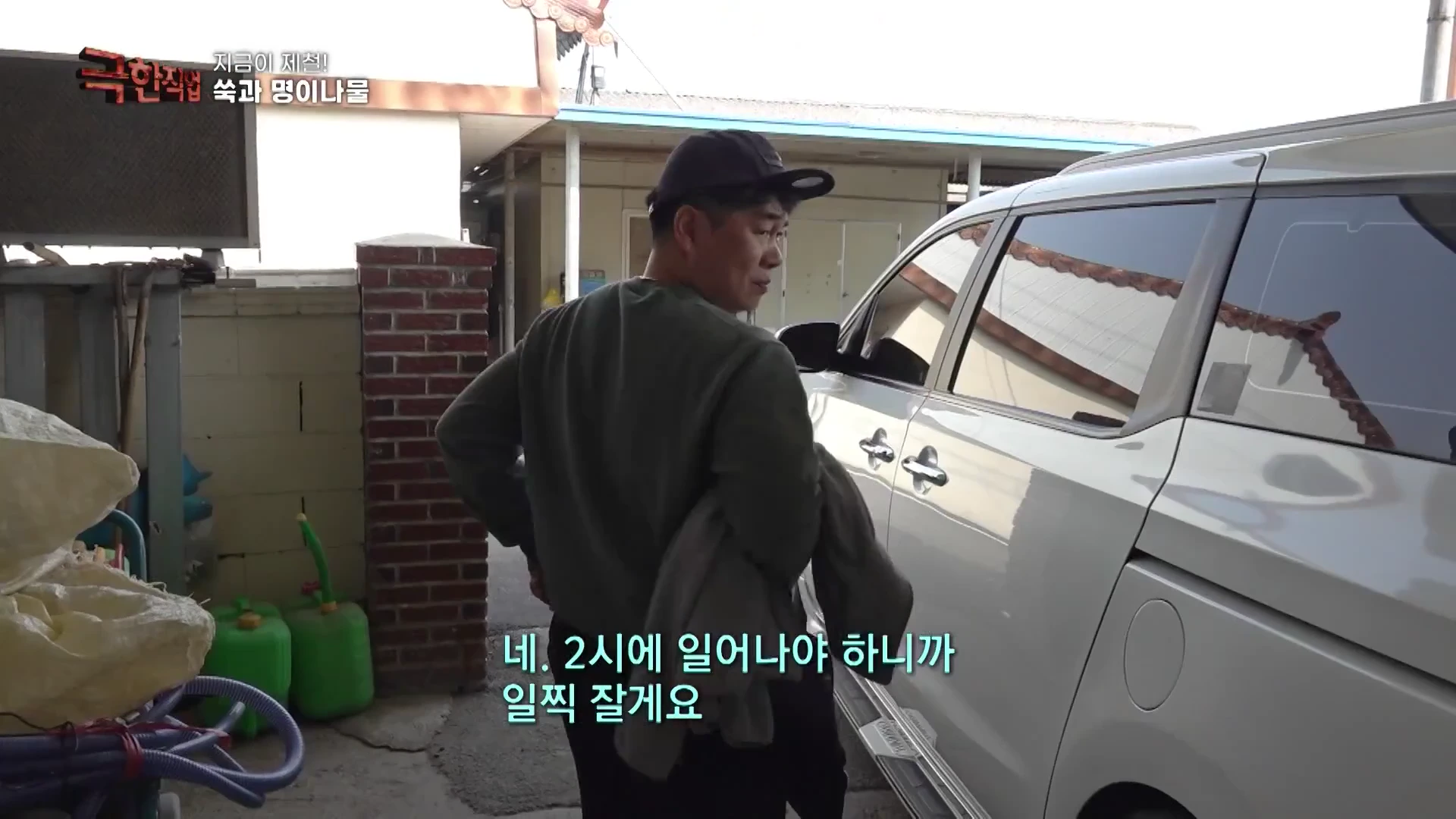 검은색 모자를 쓴 남성이 마당에 주차된 은색 승합차 옆에서 뒤를 돌아보고 있는 모습.