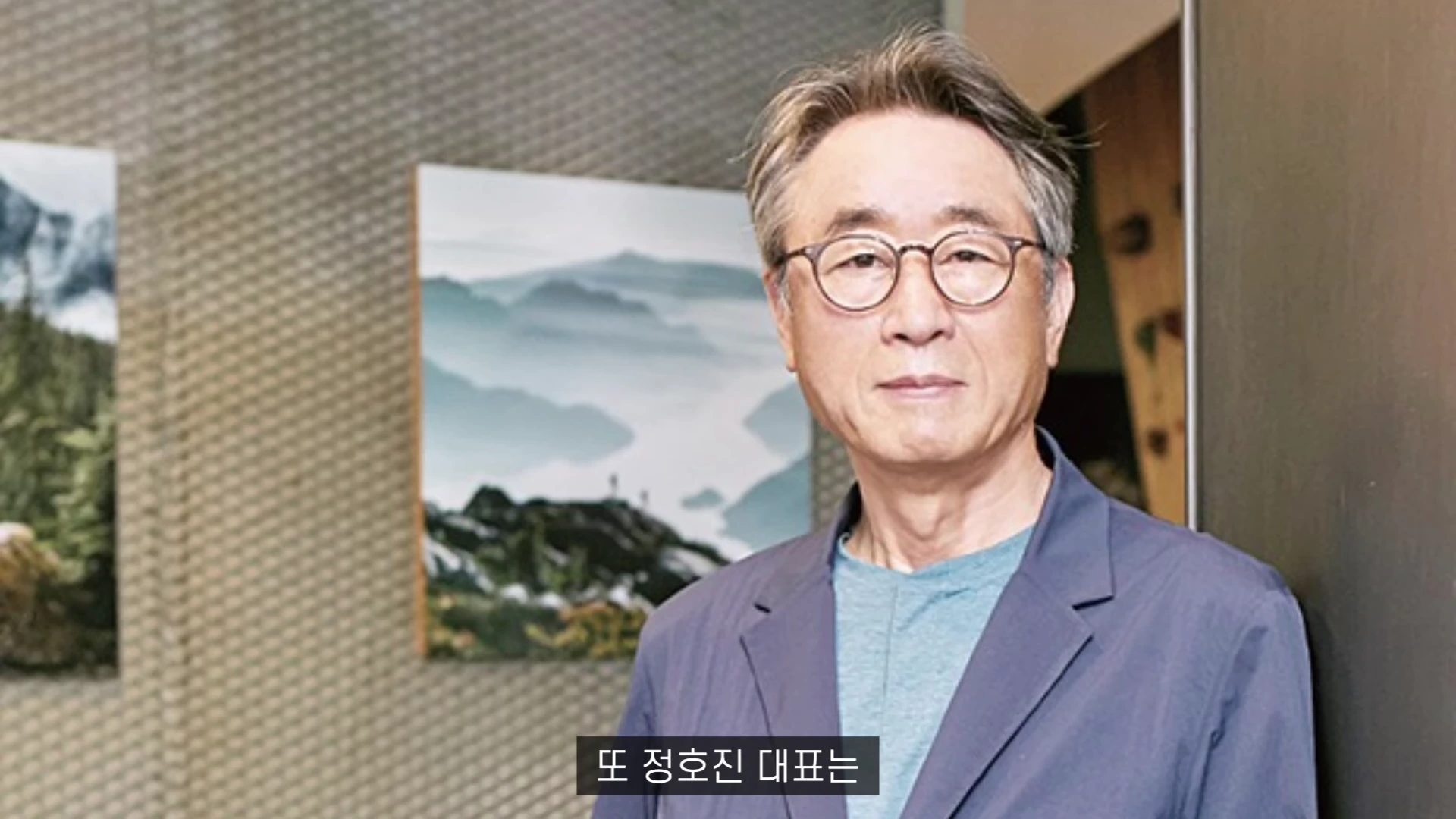 안경을 쓴 중년 남성이 회색 재킷을 입고 카메라를 정면으로 응시하고 있으며, 배경에는 산 풍경 사진이 걸려 있습니다.