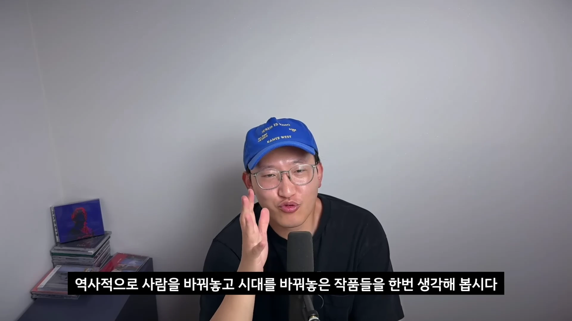 파란색 야구 모자를 쓰고 안경을 쓴 남성이 마이크 앞에 앉아 손짓하며 이야기를 나누고 있는 모습입니다. 왼쪽에는 여러 장의 CD가 쌓여 있고 화면 하단에는 자막이 떠 있습니다.