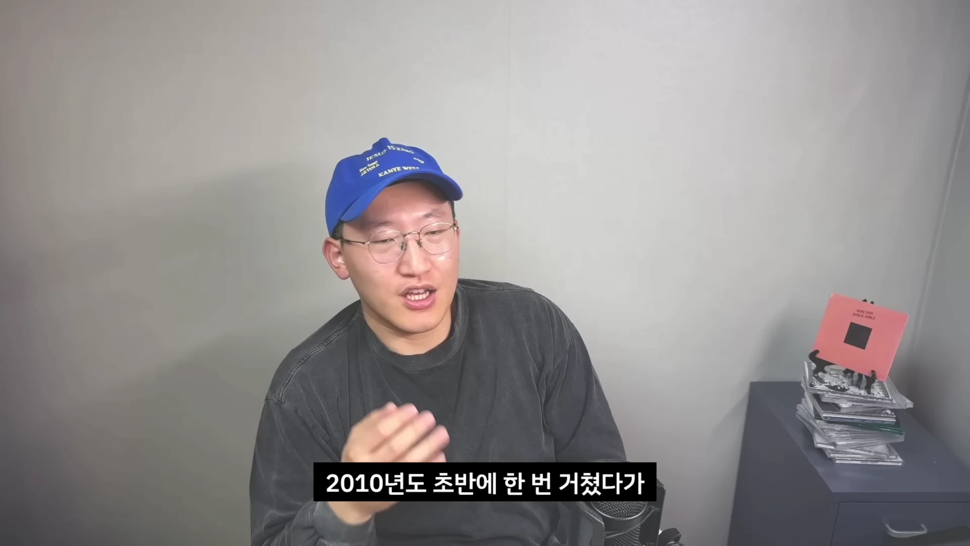 파란색 야구 모자와 안경을 쓴 남성이 무채색 배경 앞에서 정면을 바라보며 이야기하고 있다.