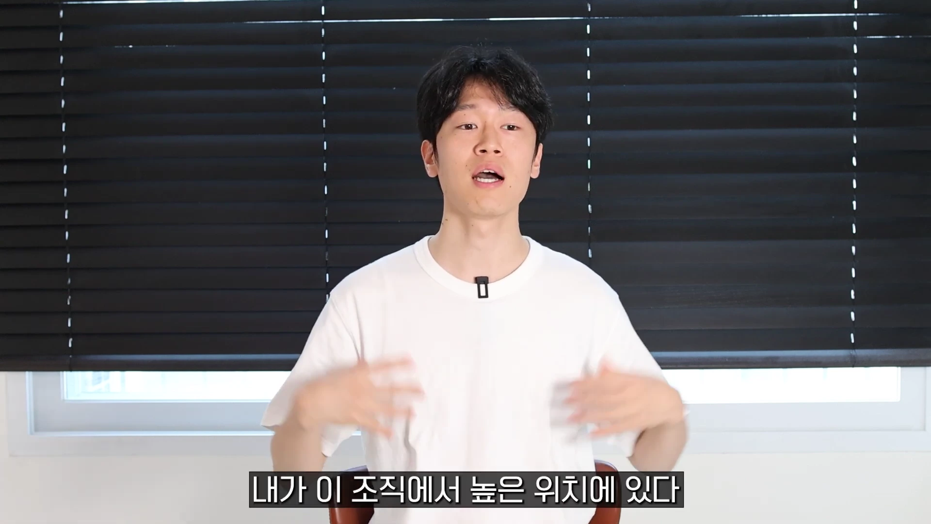 흰색 티셔츠를 입은 남성이 검은색 블라인드 배경 앞에서 손짓하며 말을 하고 있으며, 하단에는 한국어 자막이 표시되어 있습니다.
