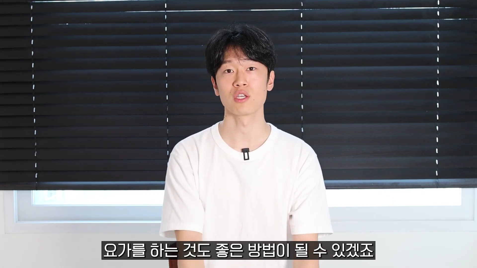 검은색 블라인드 배경 앞에 흰색 티셔츠를 입은 젊은 남성이 앉아 화면을 응시하며 이야기하고 있습니다.