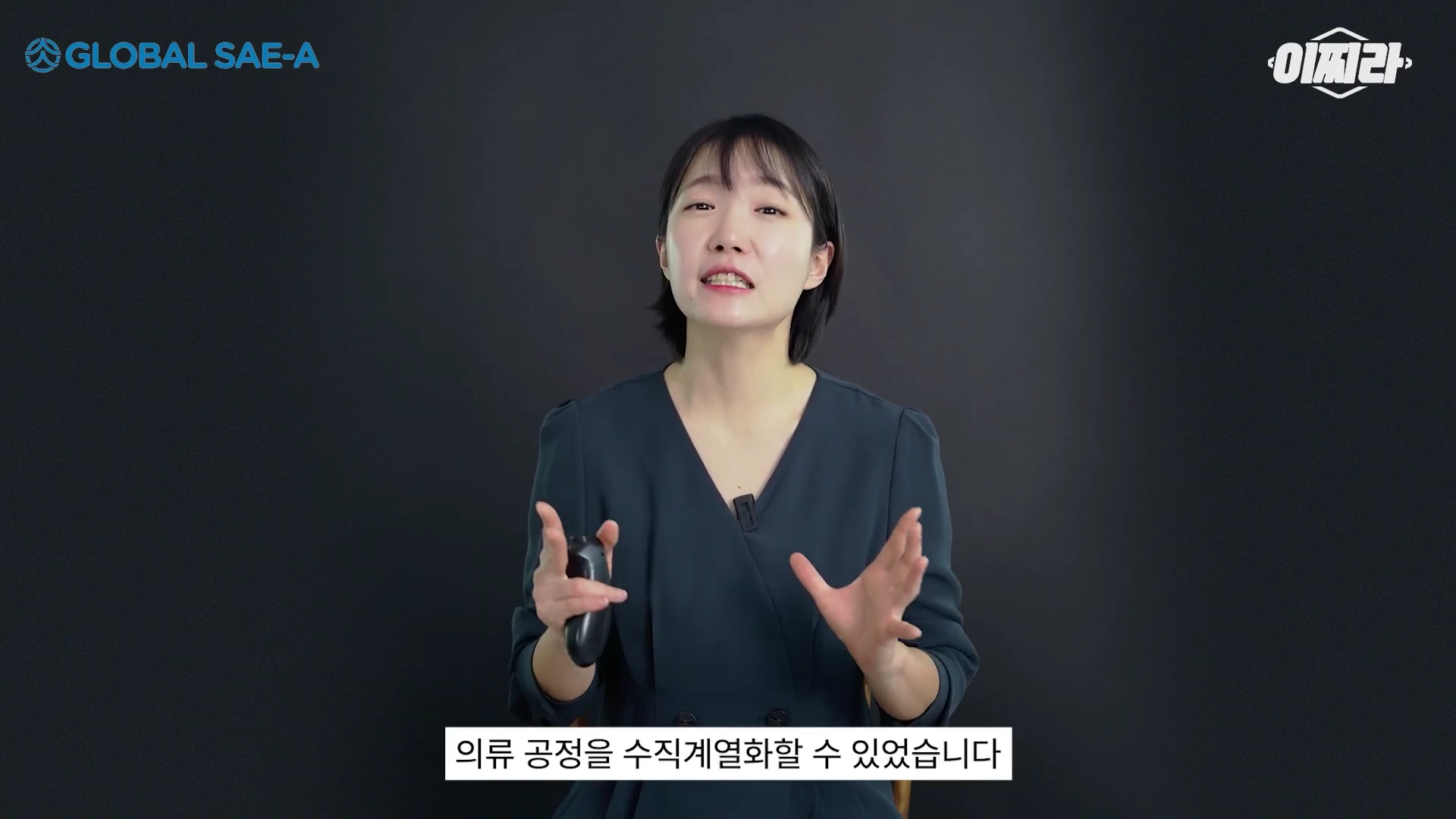 단정한 검은색 원피스를 입은 짧은 머리의 여성 발표자가 검은색 배경 앞에서 손짓을 하며 이야기를 하고 있는 스튜디오 모습입니다.