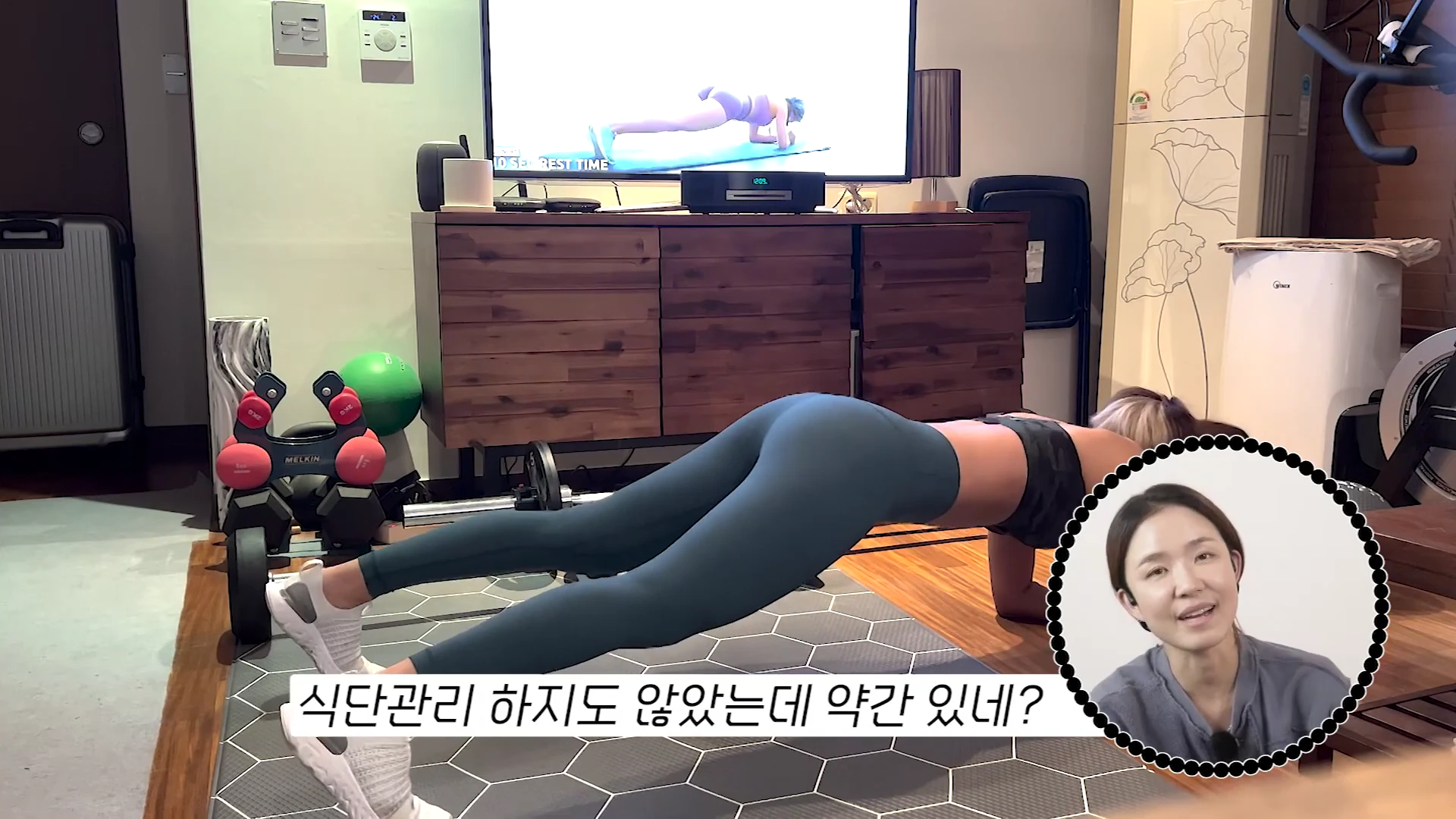 실내에서 운동복을 입은 여성이 TV 화면의 영상을 보며 플랭크 자세를 취하고 있고, 화면 우측 하단에는 말하고 있는 여성의 얼굴이 작은 원형 프레임으로 떠 있습니다.