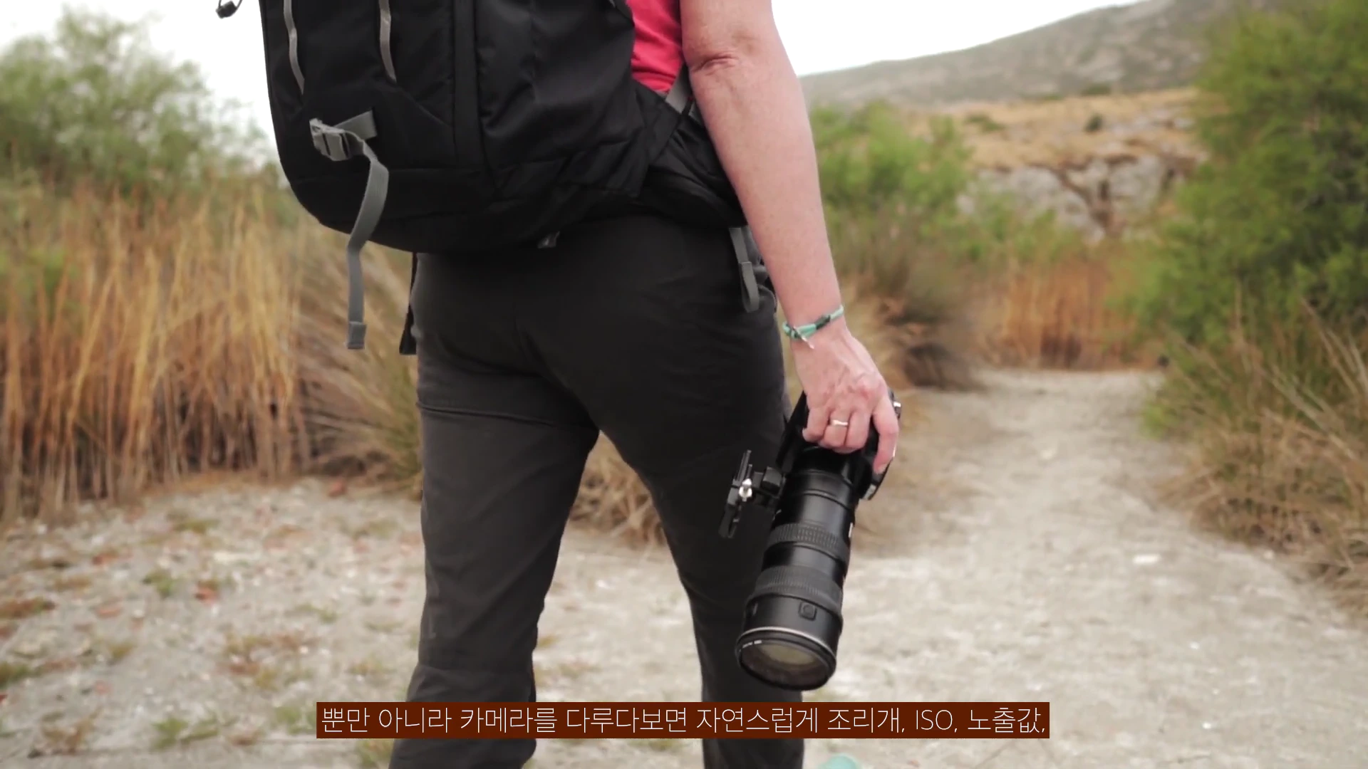배낭을 메고 카메라를 손에 든 채 야외 길을 걷고 있는 사람의 뒷모습.