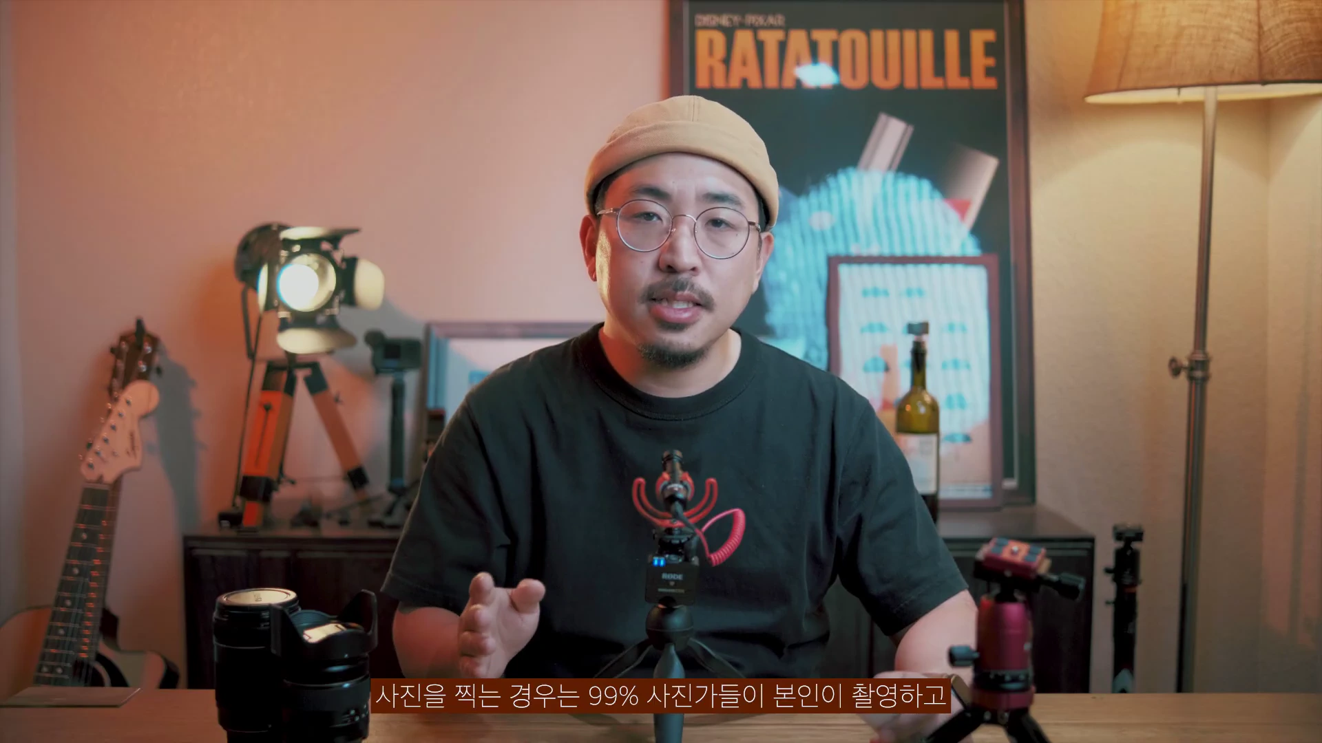안경을 쓰고 베레모를 쓴 남성이 책상에 앉아 이야기하고 있으며, 배경에는 기타와 조명, 라따뚜이 포스터가 보인다.