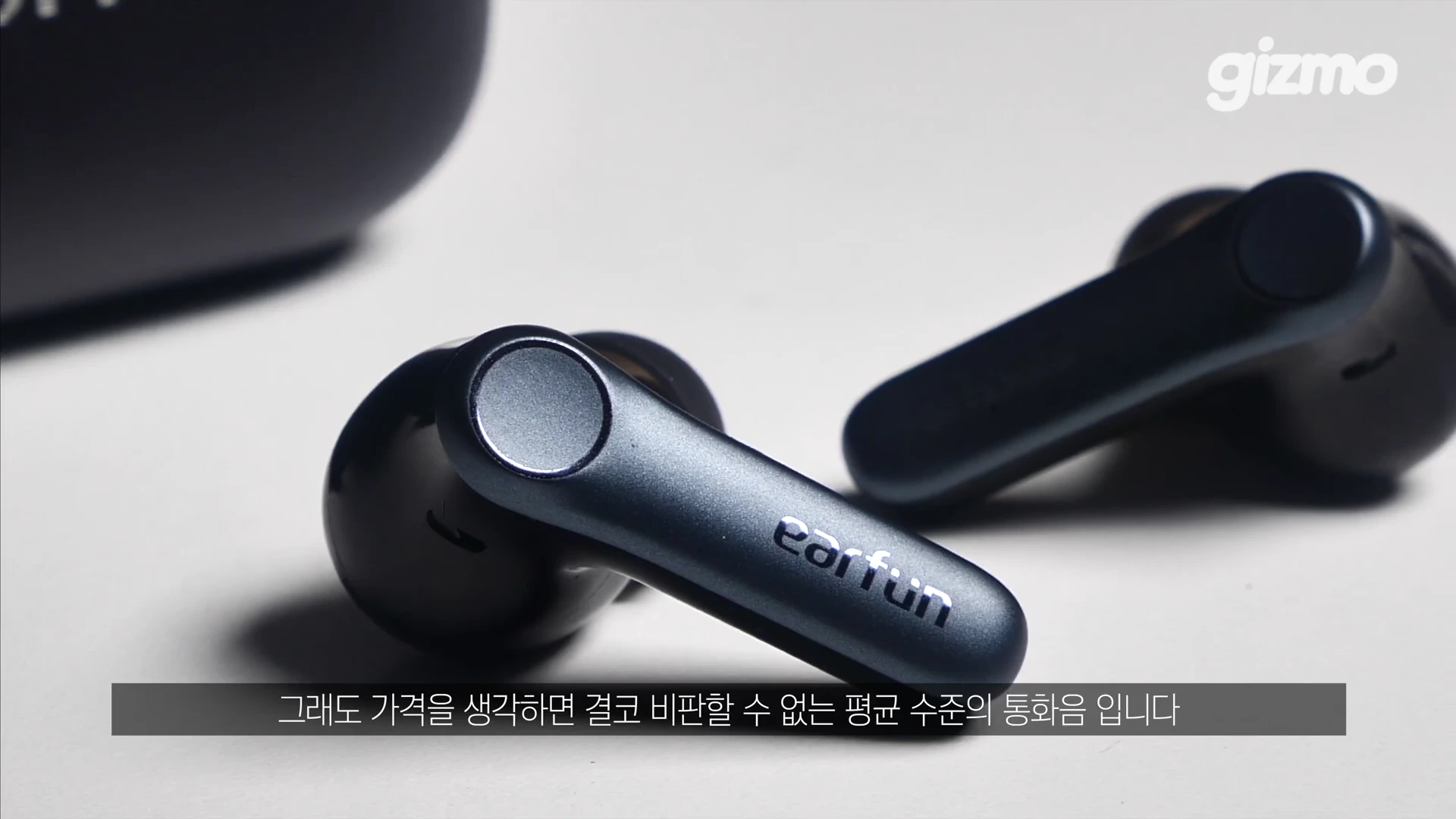 흰색 배경 위에 놓인 짙은 회색의 무선 이어폰 한 쌍과 충전 케이스 일부, 하단에는 한국어 자막이 있는 영상 캡처 화면.