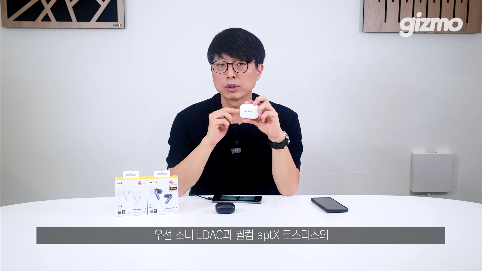 흰색 테이블 앞에 앉은 안경 쓴 남성이 이어폰 충전 케이스를 들고 설명하고 있으며, 앞에는 제품 상자와 스마트폰이 놓여 있다.