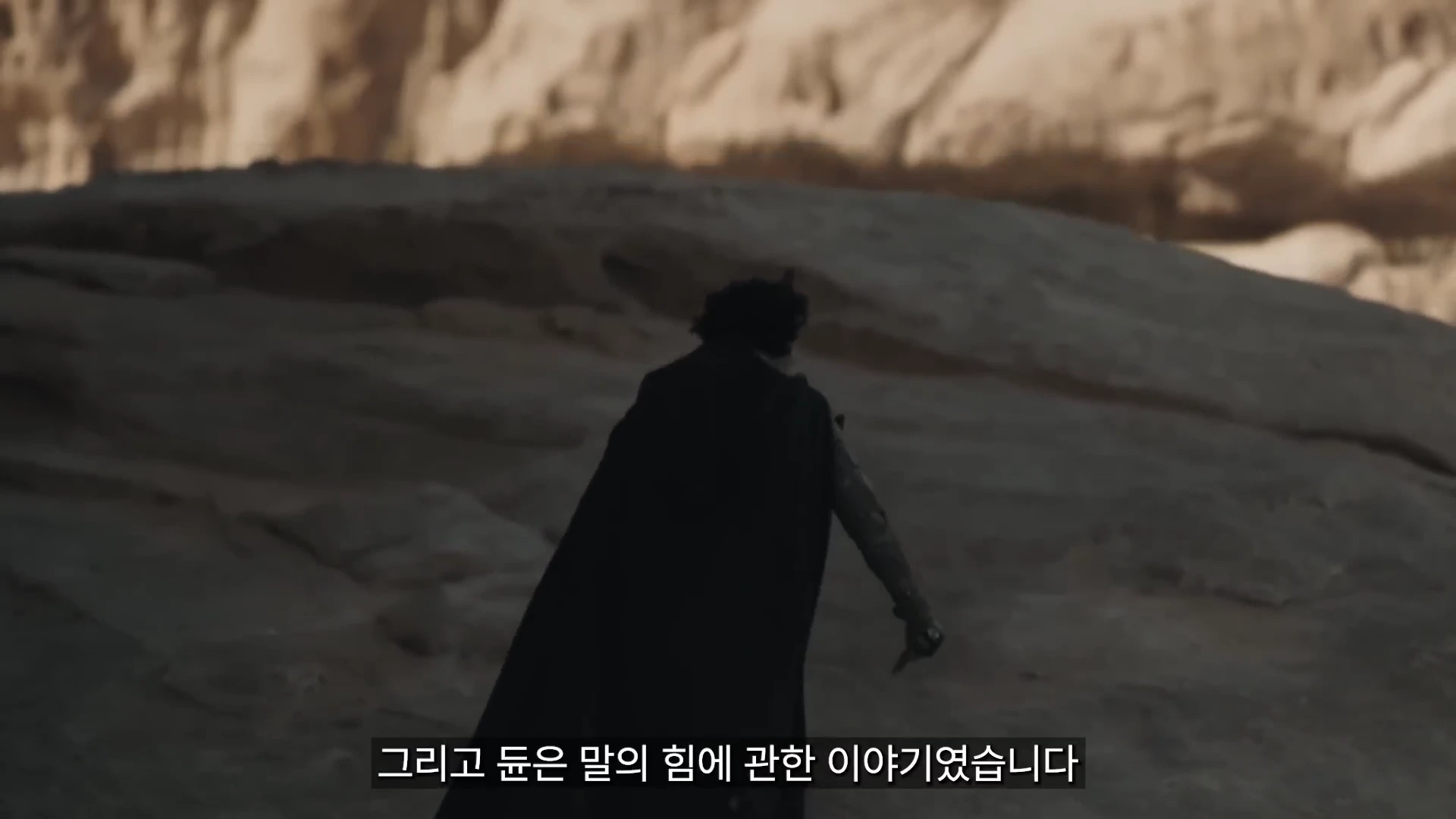 영화 속 어두운 망토를 입은 인물이 험준한 바위 지대를 걸어가고 있는 모습이며 화면 하단에는 자막이 있다.