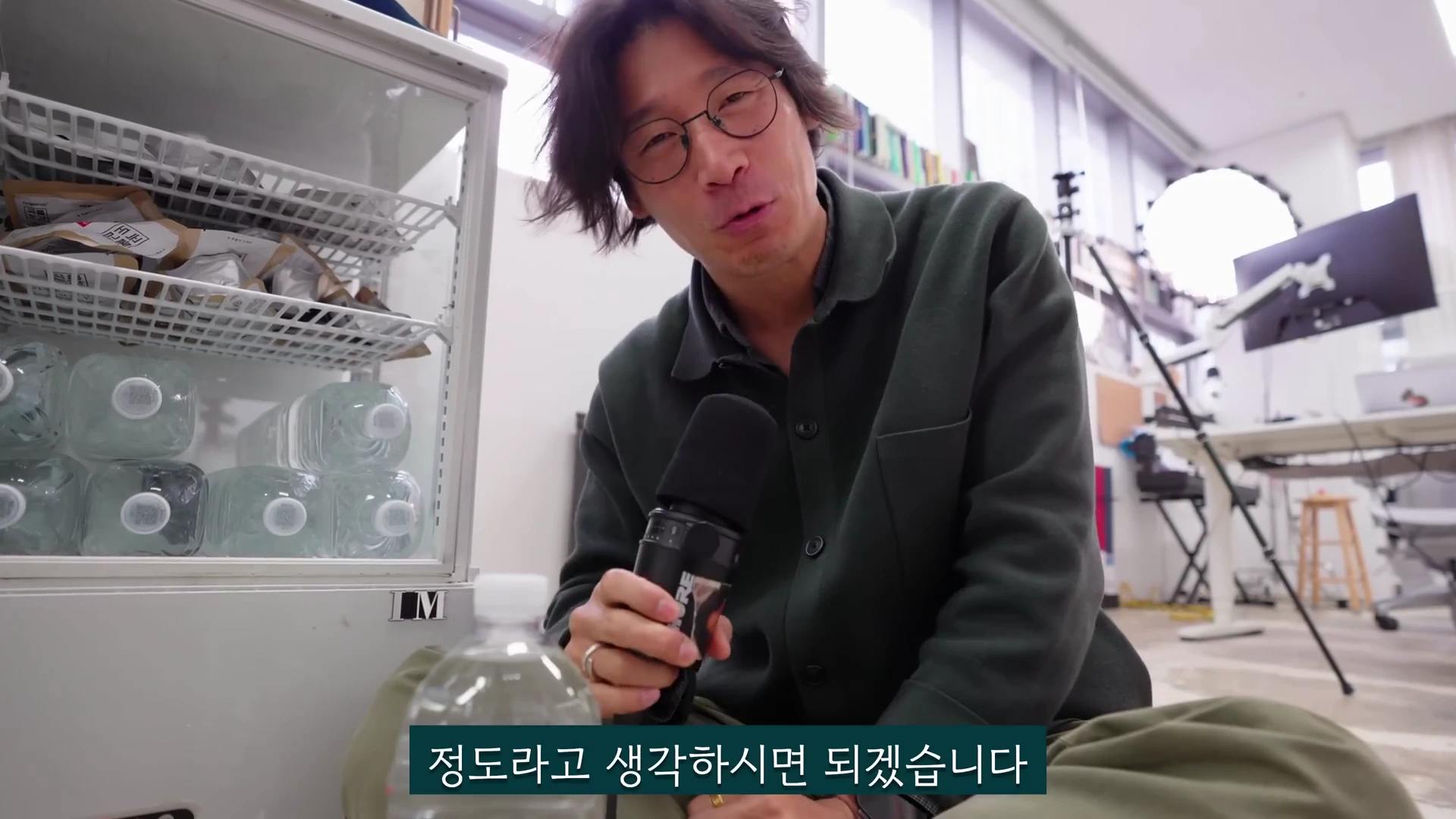 안경을 쓴 남성이 마이크를 들고 촬영용 스튜디오 내부에서 카메라를 응시하며 이야기하고 있으며, 옆에는 생수병이 가득 든 냉장고가 보임.