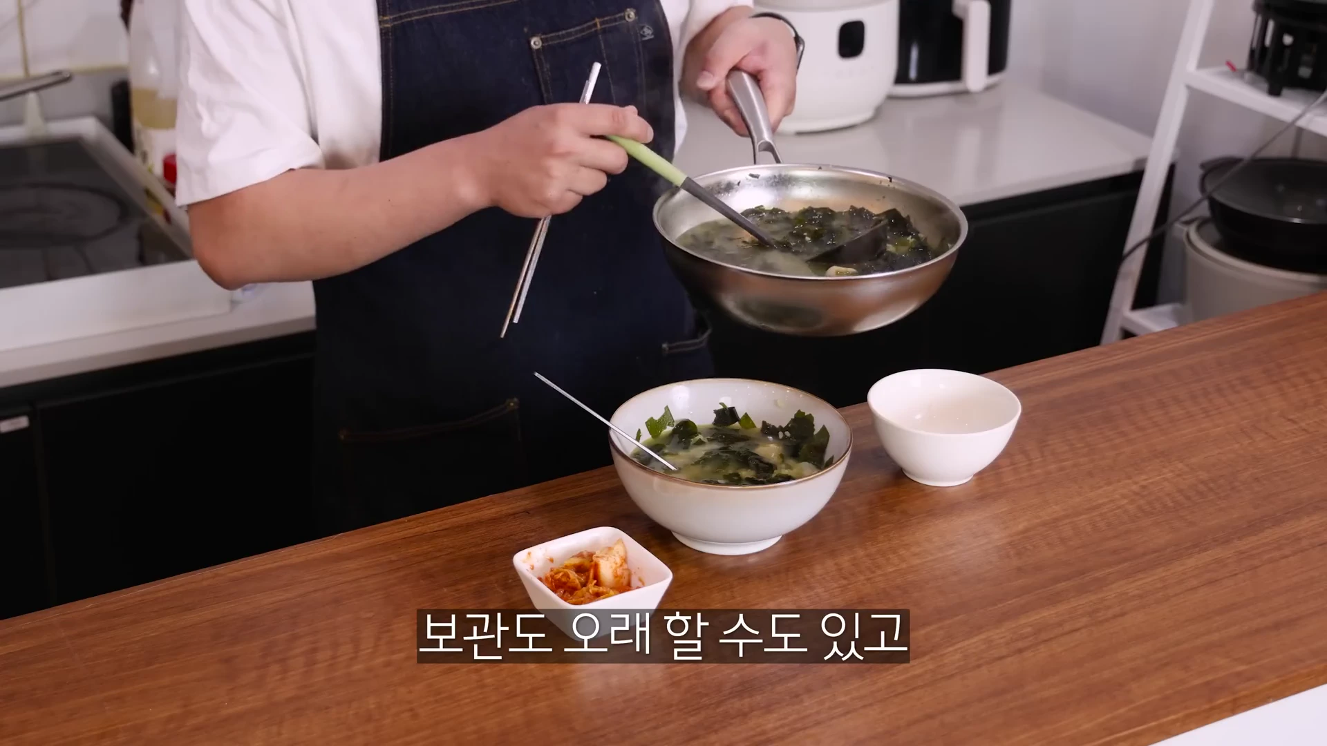앞치마를 입은 요리사가 국자로 냄비의 미역국을 그릇에 담고 있으며 식탁 위에는 완성된 미역국과 작은 그릇에 담긴 김치가 놓여 있습니다.