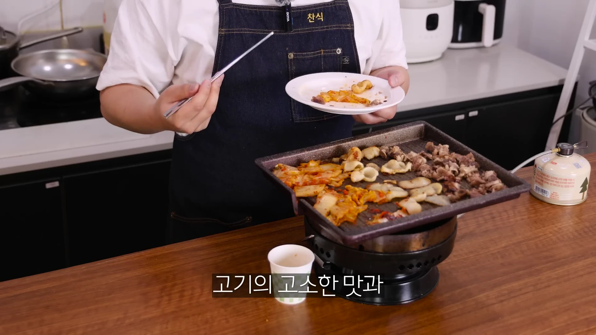 앞치마를 두른 사람이 젓가락으로 굽고 있는 관자와 김치를 앞접시에 옮겨 담고 있는 모습