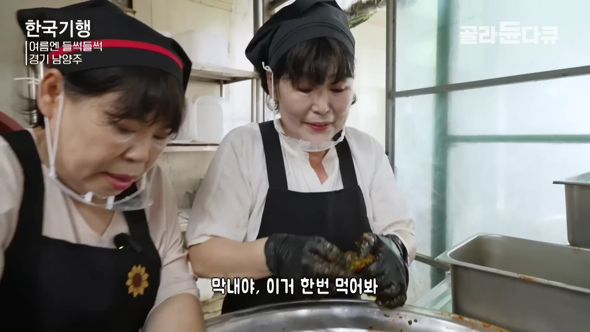 두 명의 중년 여성이 주방에서 검은색 앞치마와 위생 모자를 쓰고 오이소박이를 담그고 있는 모습이다.
