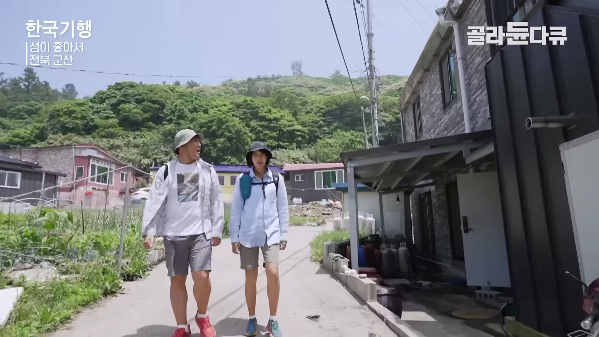 두 명의 여행자가 모자를 쓰고 배낭을 멘 채 햇살이 비치는 작은 섬 마을 길을 나란히 걷고 있다.