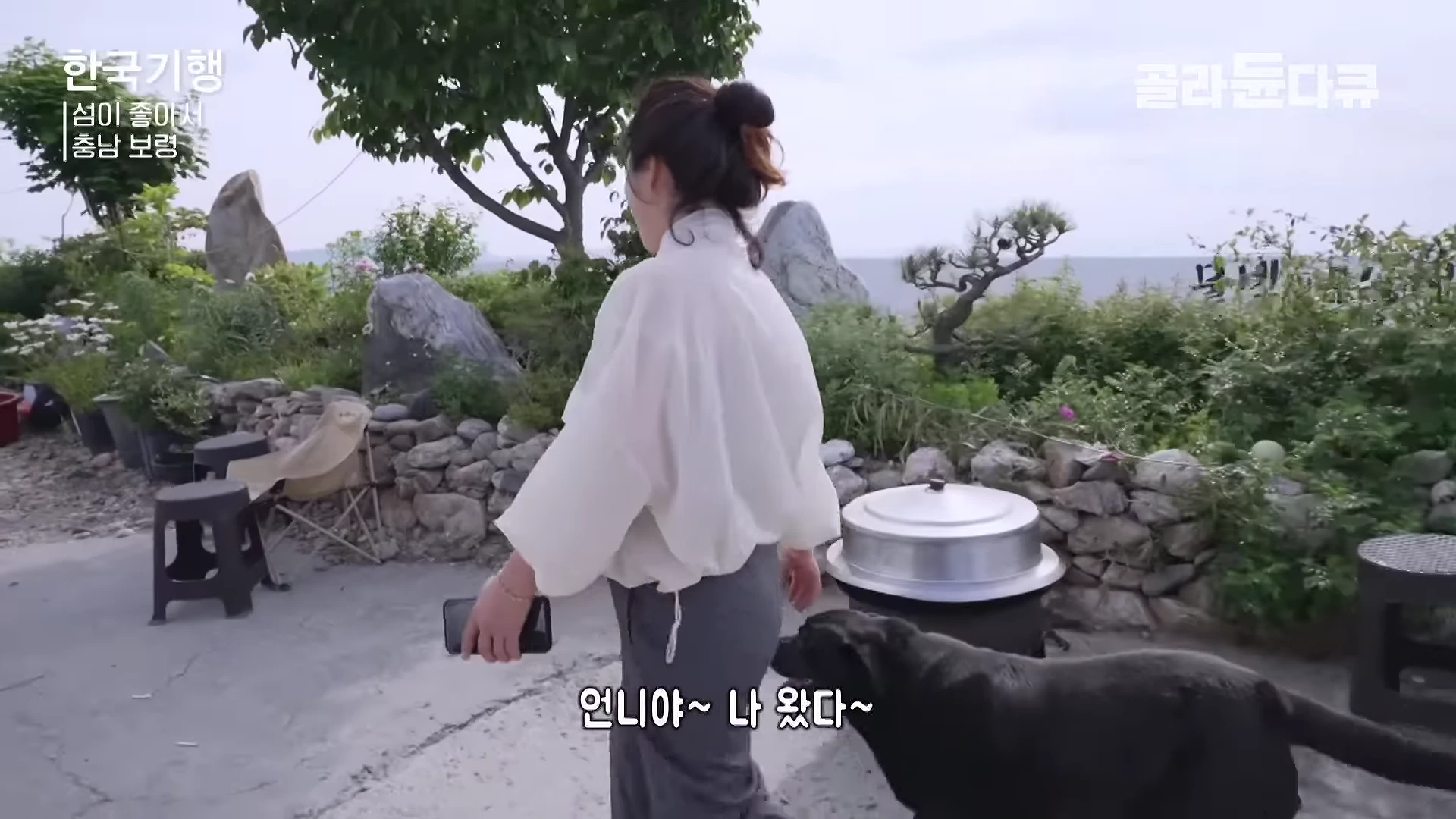 흰 셔츠를 입고 머리를 묶은 여성이 강아지와 함께 바다가 보이는 정원으로 걸어가는 뒷모습을 담은 영상 장면입니다.