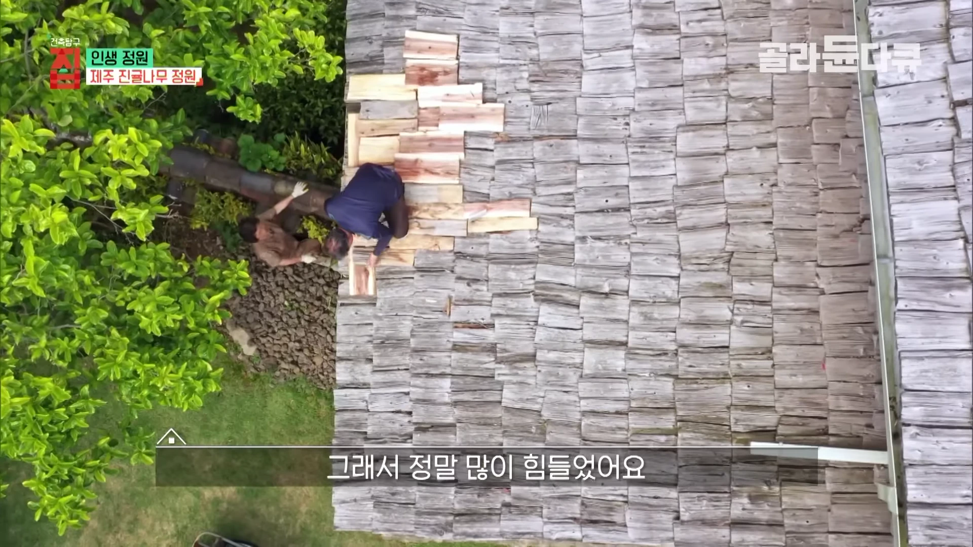 항공에서 내려다본 지붕 수리 현장으로, 나무 판자를 덧대어 지붕을 보수하는 두 사람과 주변의 푸른 정원이 보입니다.