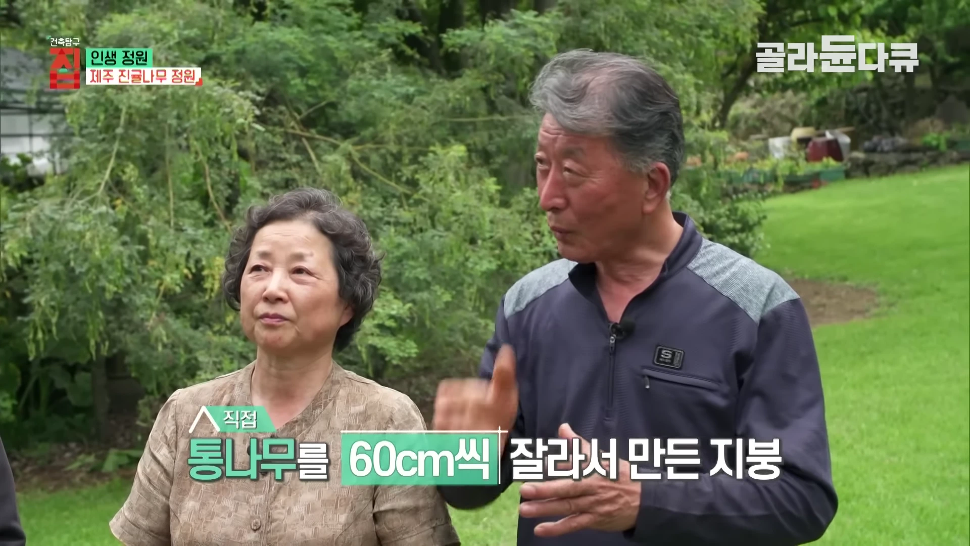An elderly couple standing in a garden, with the man speaking and gesturing with his hand and the woman listening beside him, with text overlays on the screen.