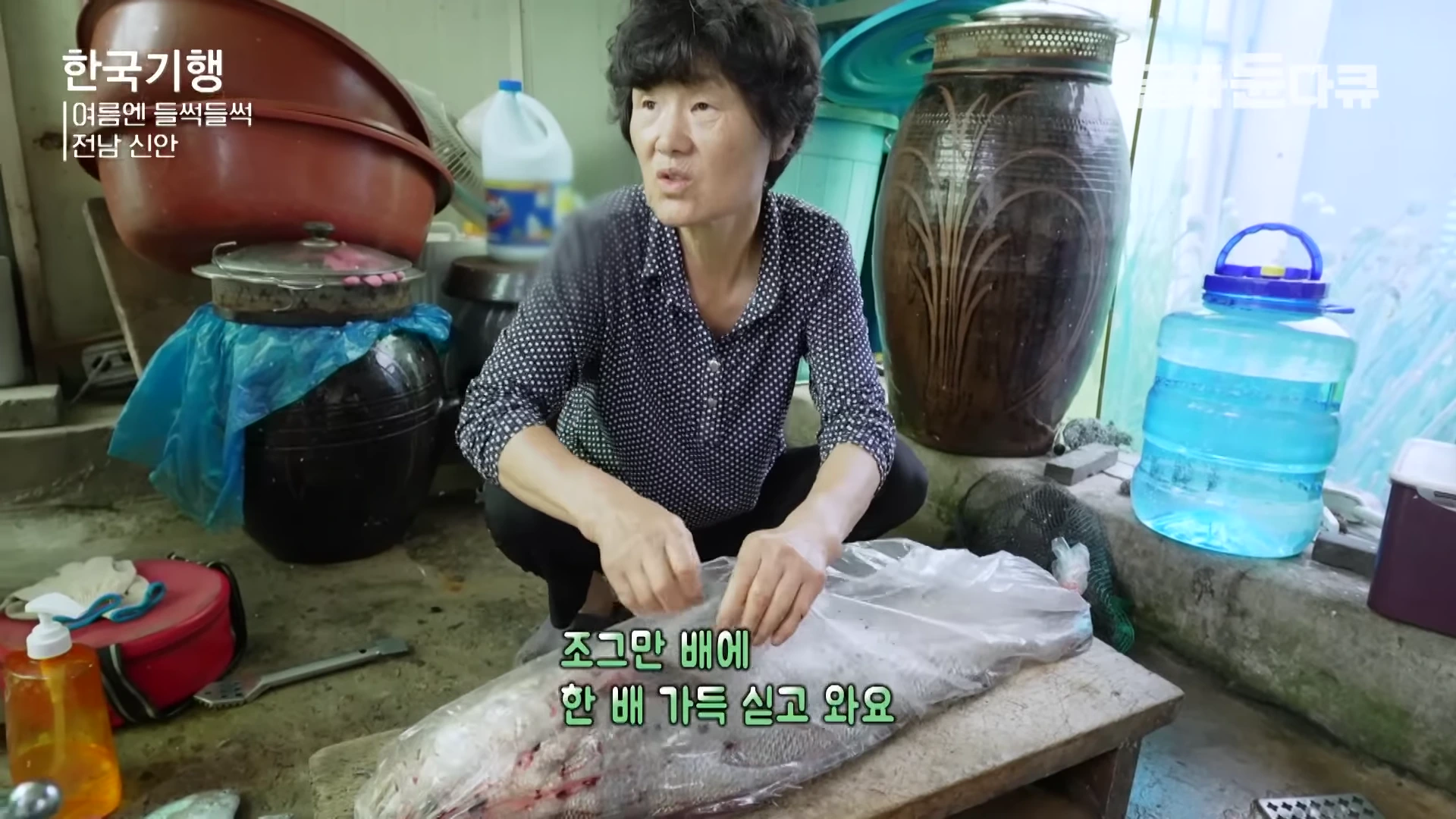 실내에서 중년 여성이 비닐에 싸인 큰 생선을 앞에 두고 앉아 있는 모습.