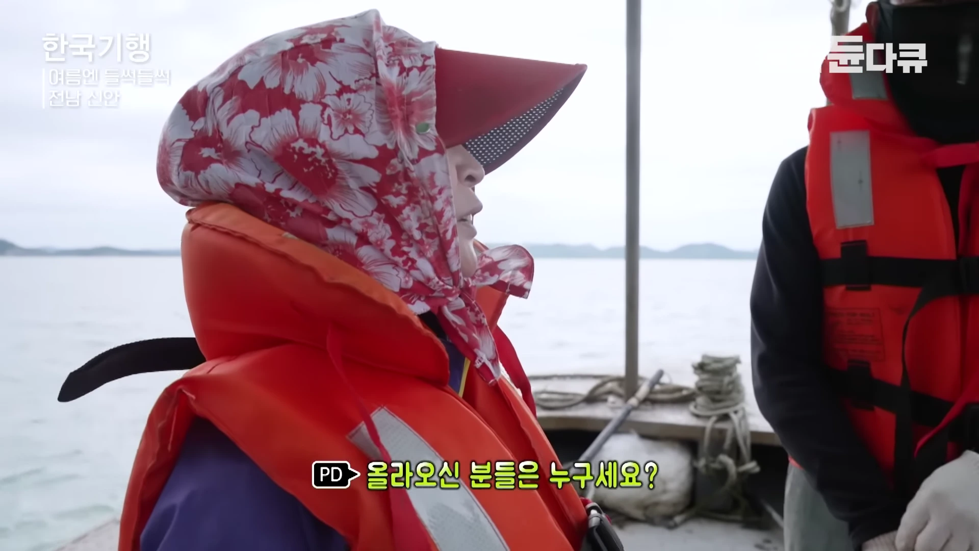 꽃무늬 두건과 빨간 챙 모자를 쓴 중년 여성이 오렌지색 구명조끼를 입고 배 위에서 누군가와 대화하고 있는 모습