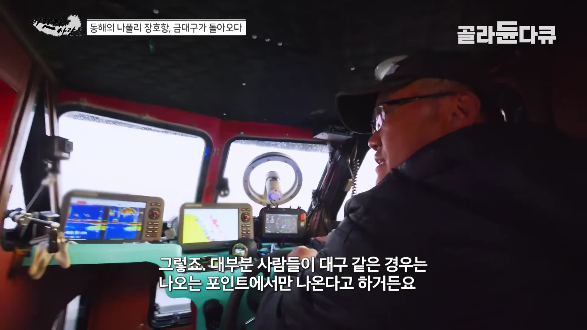 선박 조타실 안에서 모자와 안경을 쓴 남성이 여러 대의 항해용 모니터와 장비를 보며 대화하고 있다.