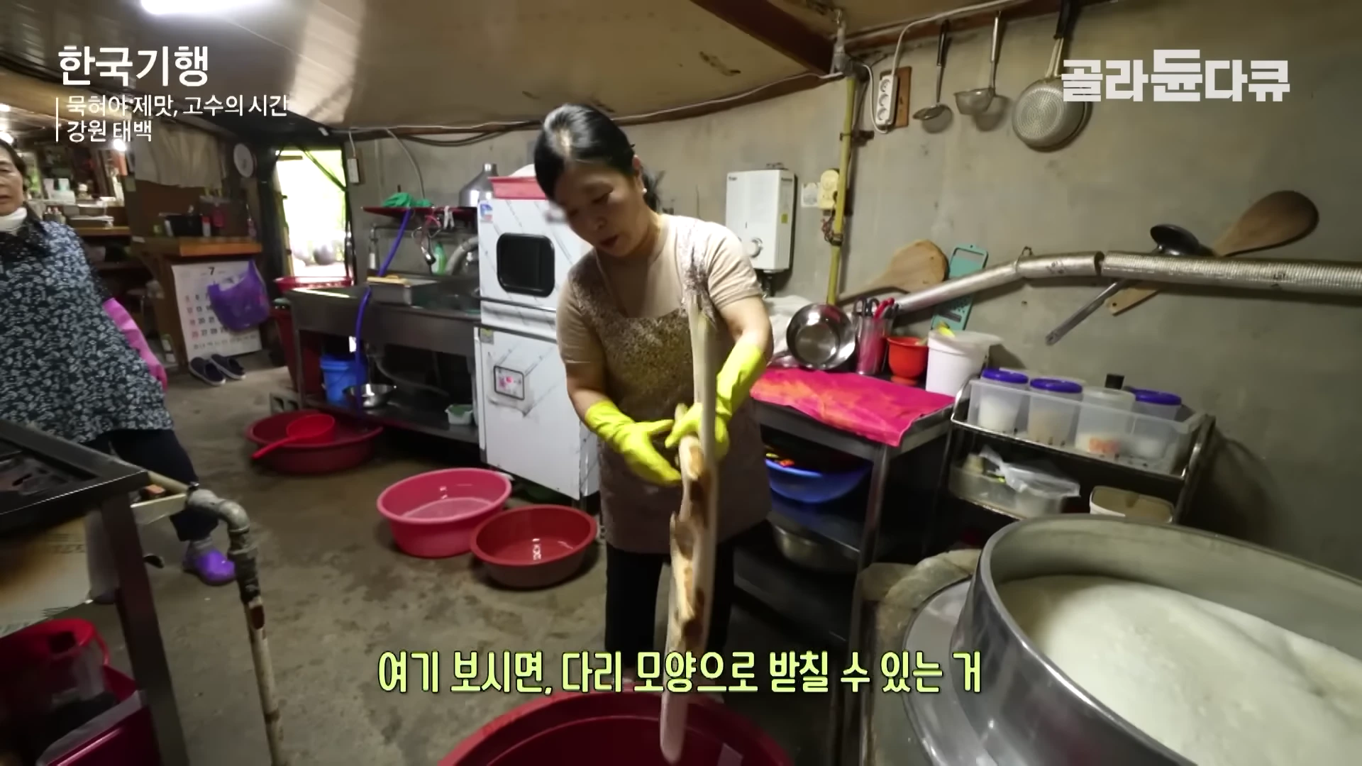 주방에서 노란 고무장갑을 낀 여성이 오래된 나무 도구로 순두부를 만들고 있고, 옆에는 다른 여성이 서 있는 모습