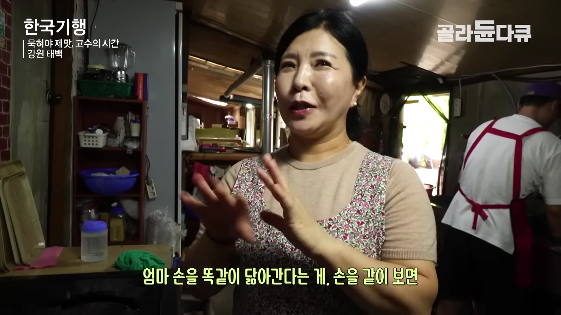 꽃무늬 앞치마를 입은 중년 여성이 주방에서 손짓하며 이야기를 나누고 있고, 뒤편에는 흰 상의에 빨간 앞치마를 두른 남성이 작업하고 있는 모습입니다.