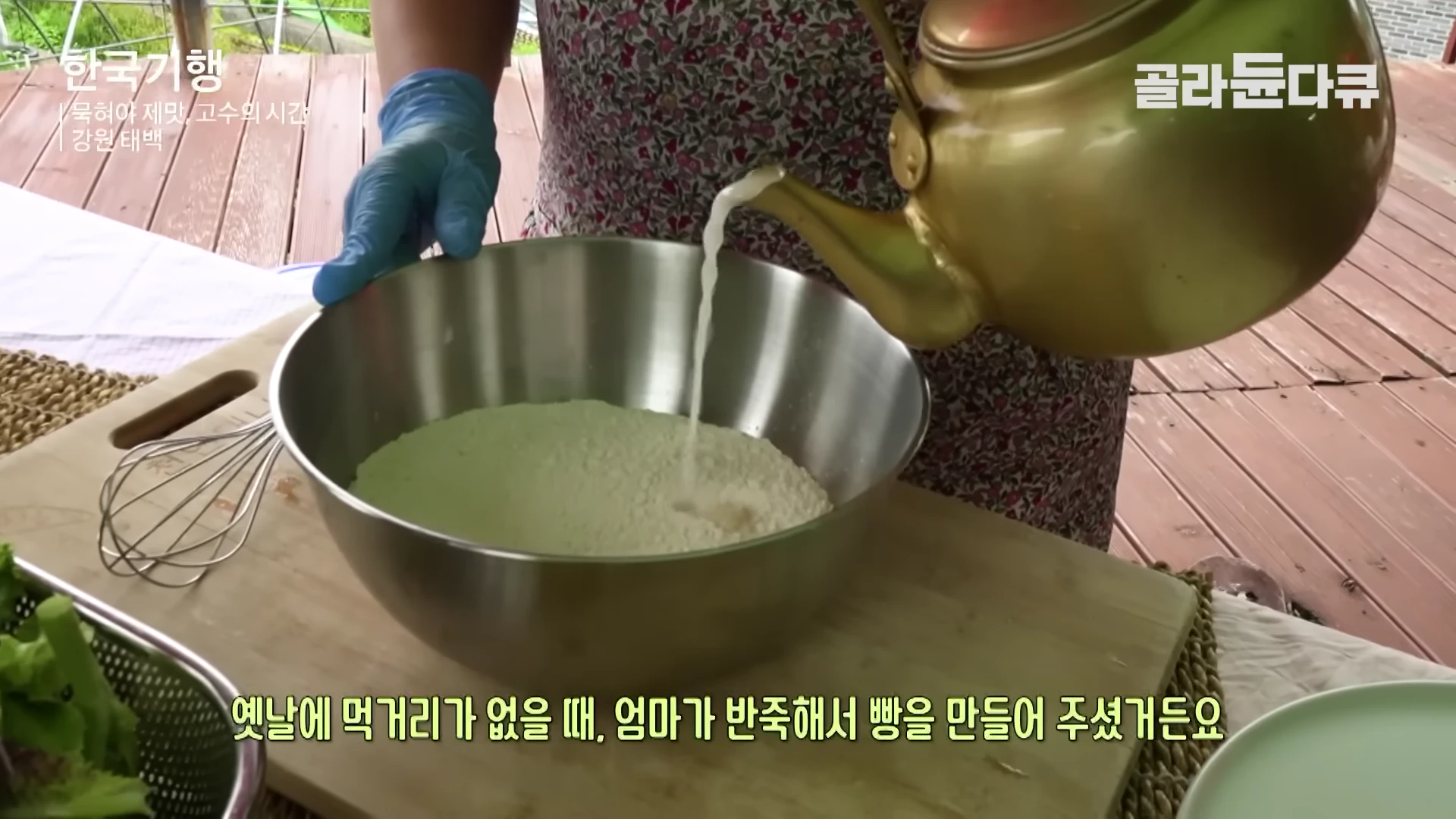 파란색 고무장갑을 낀 손이 노란색 주전자로 스테인리스 볼에 담긴 밀가루 반죽에 막걸리를 붓고 있다.