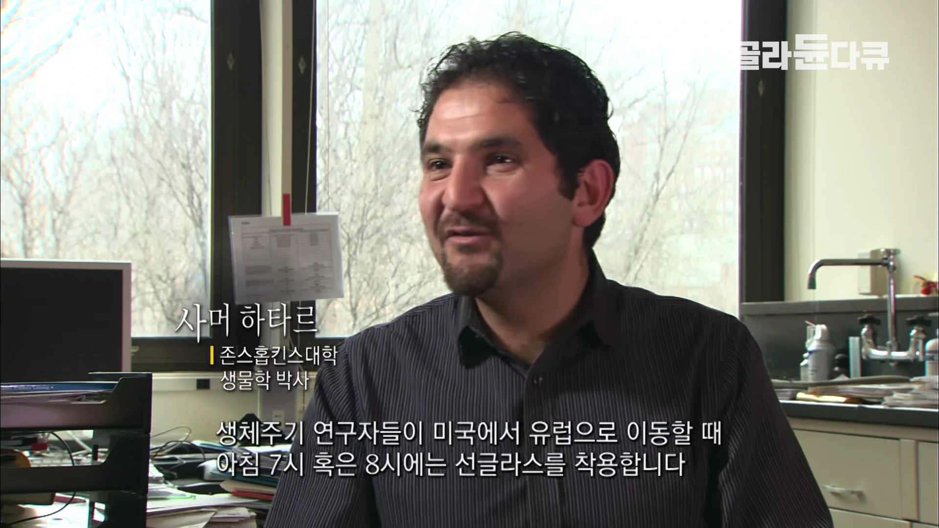실내에서 검은색 줄무늬 셔츠를 입은 남성이 인터뷰에 응하고 있으며, 하단에는 이름과 소속이 자막으로 표시되어 있습니다.
