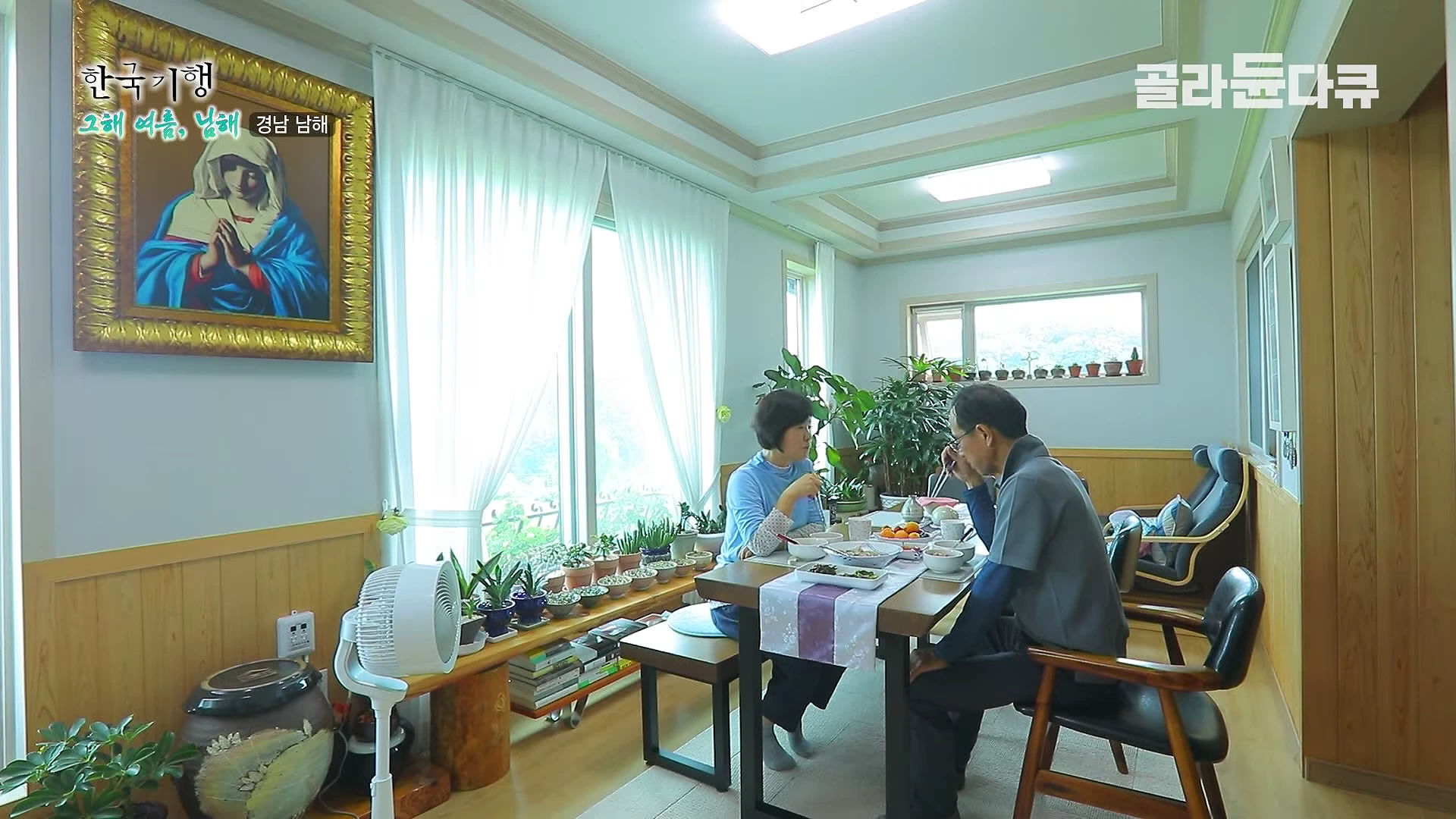 밝은 채광이 들어오는 깔끔한 거실 식탁에서 중년 부부가 함께 식사를 하고 있으며, 벽면에는 성모 마리아 그림이 걸려 있다.