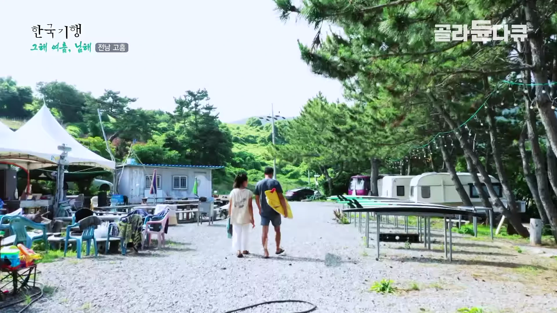 서핑 보드를 든 남성과 흰 옷을 입은 여성이 울창한 나무가 있는 바닷가 캠핑장 길을 나란히 걸어가고 있다.