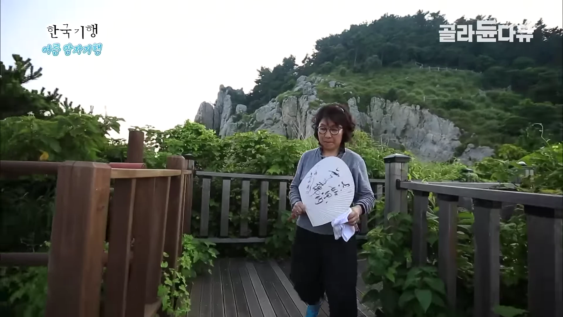 안경을 쓴 여성이 암자 데크 위에서 부채를 들고 걸어오는 모습이며 뒤편으로 거대한 바위산이 펼쳐져 있다.