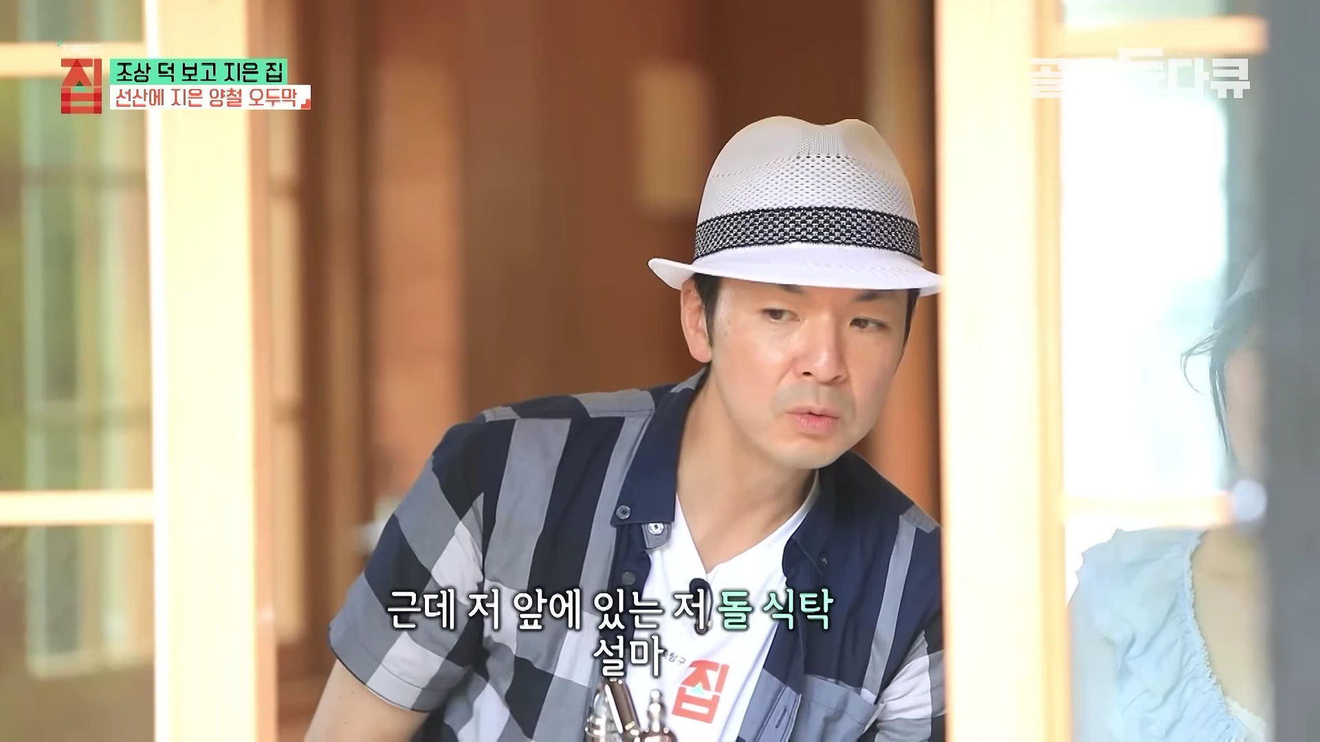 중절모를 쓴 남성이 실내에서 창밖을 바라보며 대화하고 있는 모습