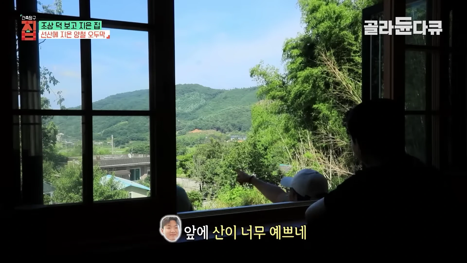 실내에서 창문을 통해 바라본 푸른 숲과 산의 풍경, 그 앞에 앉아 있는 부부의 뒷모습.