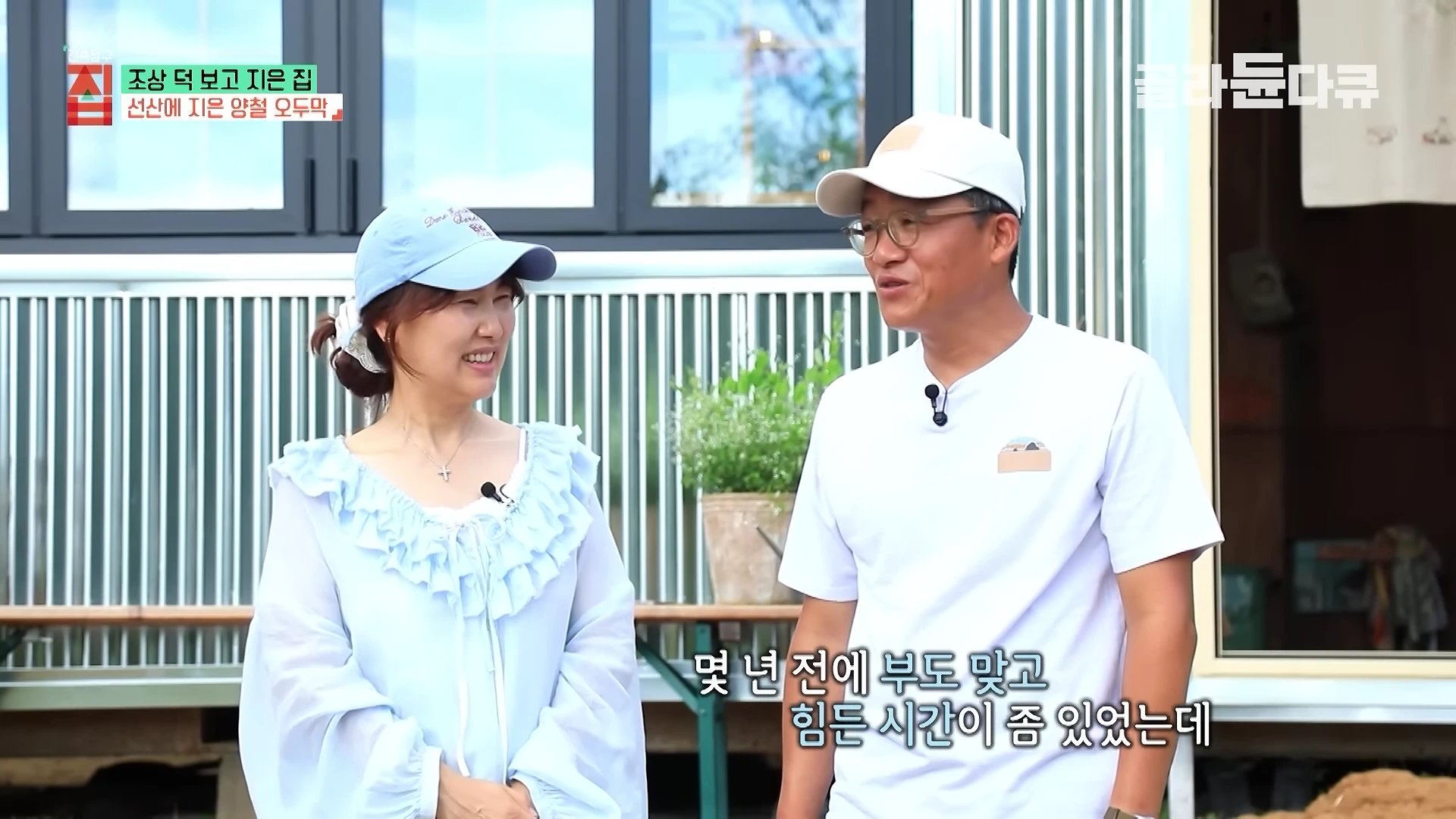 야외에서 밝게 웃으며 대화를 나누는 중년 부부의 모습으로 뒤편에는 양철 벽면의 주택 일부가 보인다.