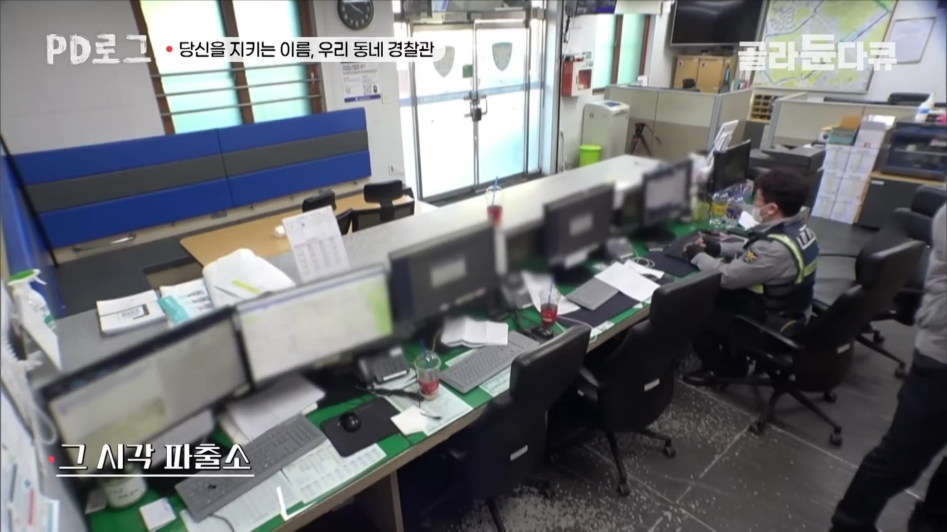 파출소 내부 사무 공간을 위에서 내려다본 모습으로 경찰관 한 명이 업무용 컴퓨터 앞에 앉아 있고 여러 대의 모니터와 서류가 책상 위에 놓여 있다.