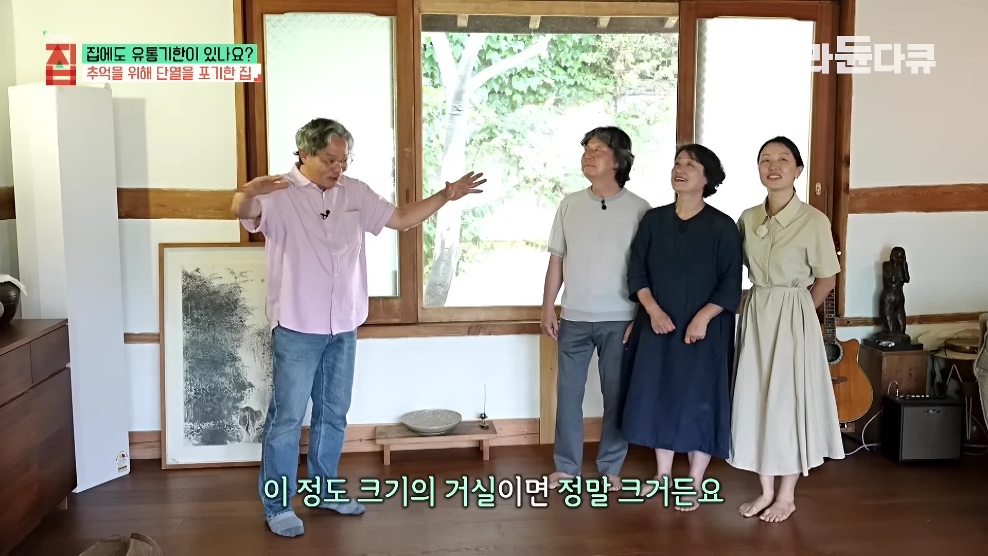 실내 거실에 서 있는 네 사람 중 한 남성이 손을 벌려 거실의 너비를 설명하고 있으며 뒤편에는 나무 창틀과 유리창이 보인다.