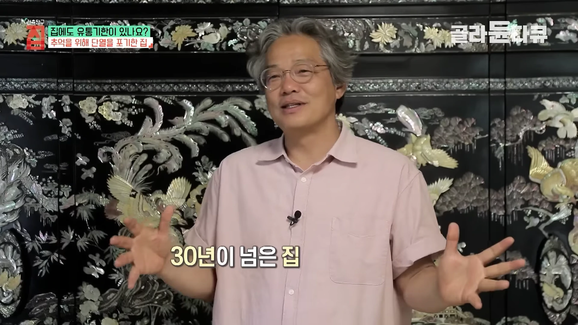 실내에서 분홍색 셔츠를 입은 중년 남성이 화려한 자개장 앞에 서서 손짓하며 이야기를 나누고 있다.