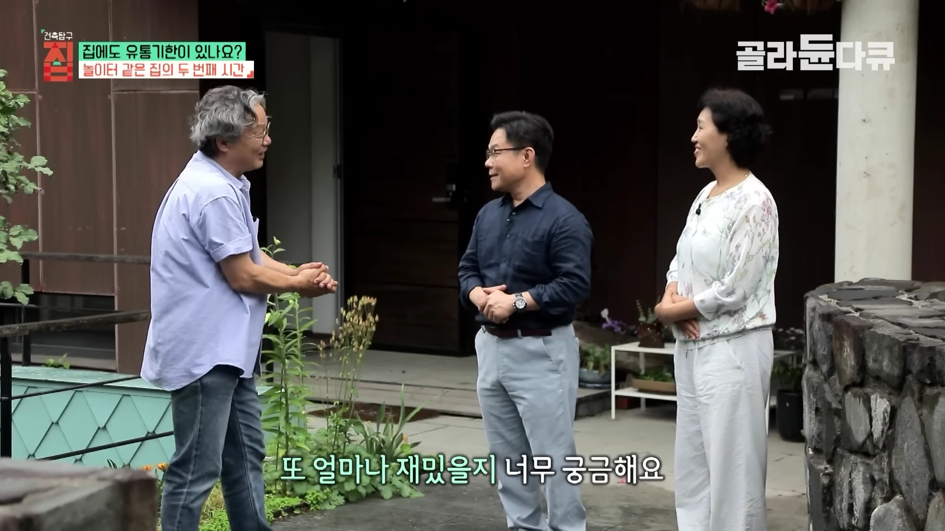 두 남성과 한 여성이 야외에서 서로 마주 보며 대화를 나누고 있는 모습. 배경에는 나무와 돌로 지어진 독특한 형태의 주택 입구가 보입니다.