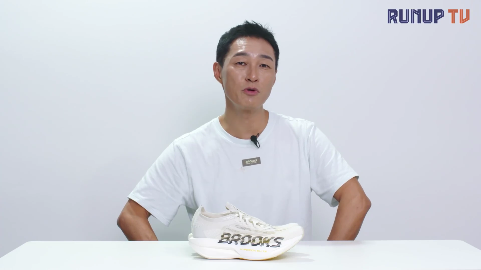 흰색 티셔츠를 입은 남성이 흰색 테이블 위에 놓인 브룩스 러닝화를 손으로 가리키며 설명하고 있다.