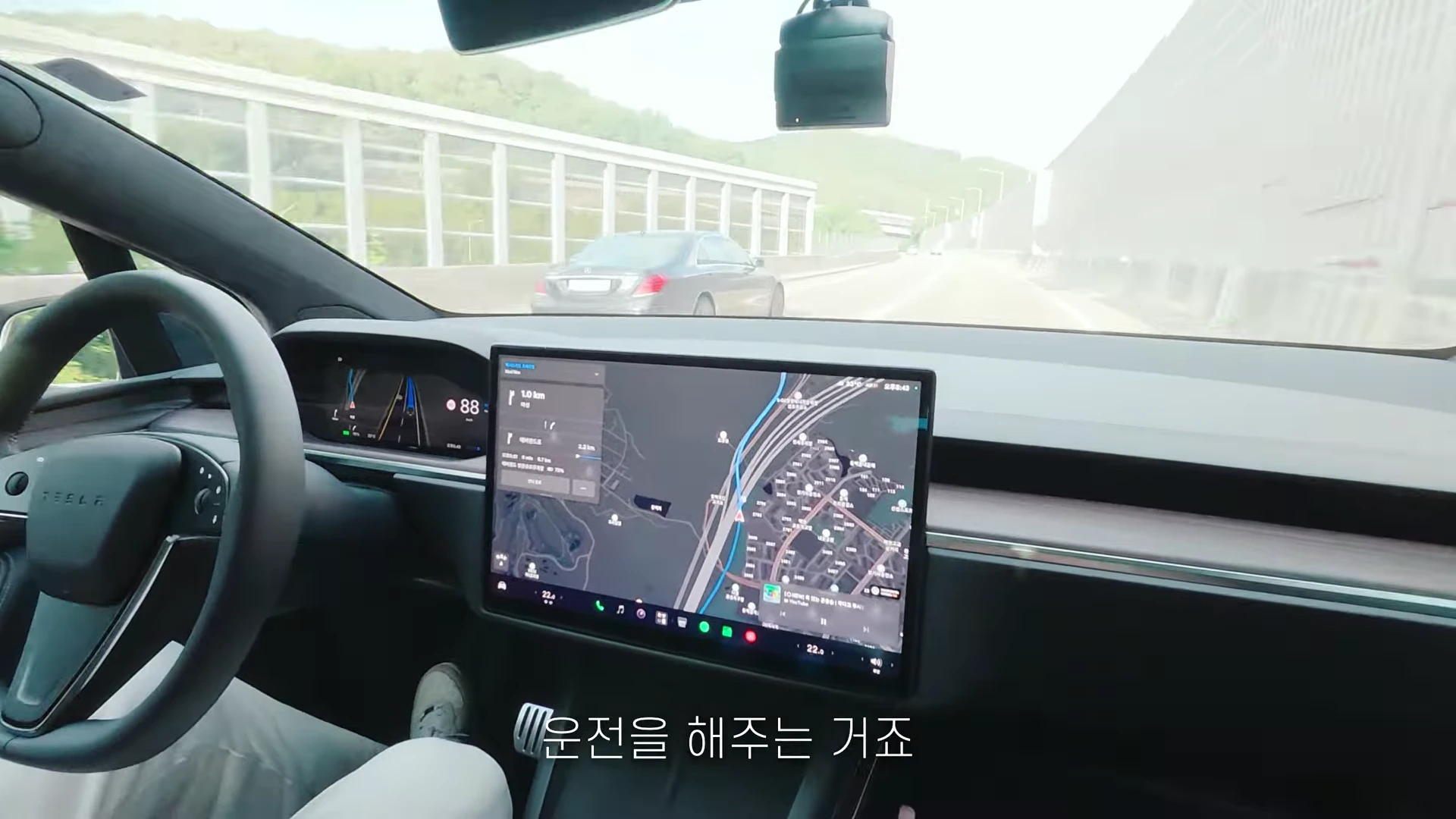테슬라 차량 내부에서 바라본 도로 주행 상황으로 운전석 스티어링 휠과 내비게이션 화면이 보이며 앞 유리창을 통해 고속도로가 펼쳐져 있다.