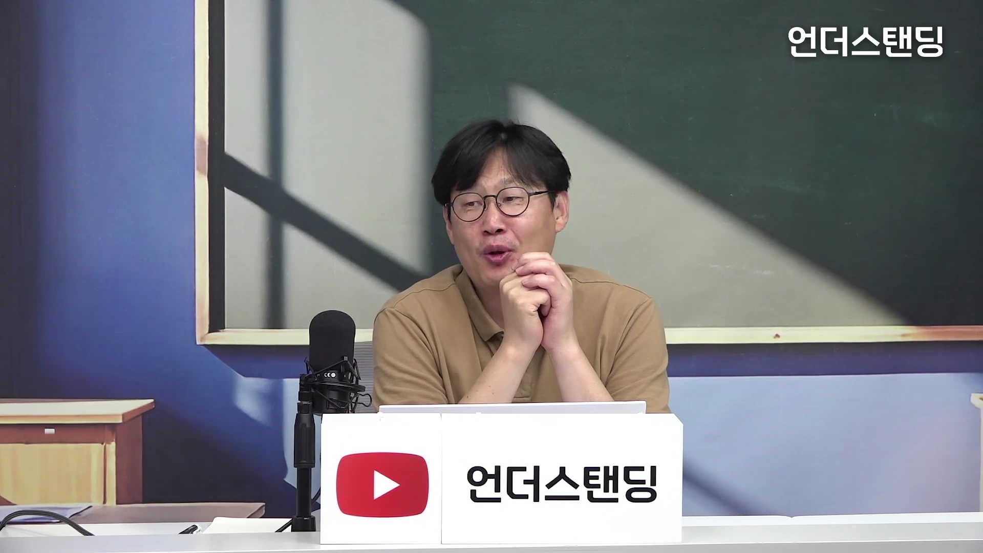 안경을 쓴 남성이 스튜디오 책상 앞에 앉아 양손을 모은 채 이야기를 나누고 있다.