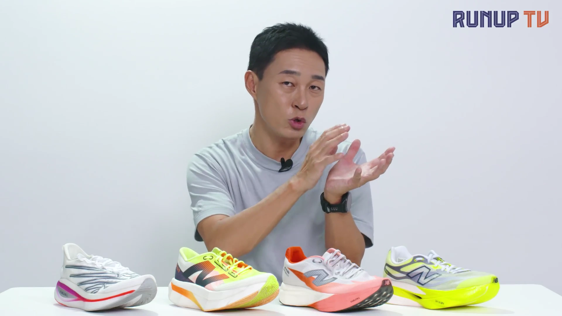 흰색 배경의 스튜디오에서 남성이 책상 위에 놓인 네 켤레의 러닝화를 앞에 두고 손동작을 하며 설명하고 있다.